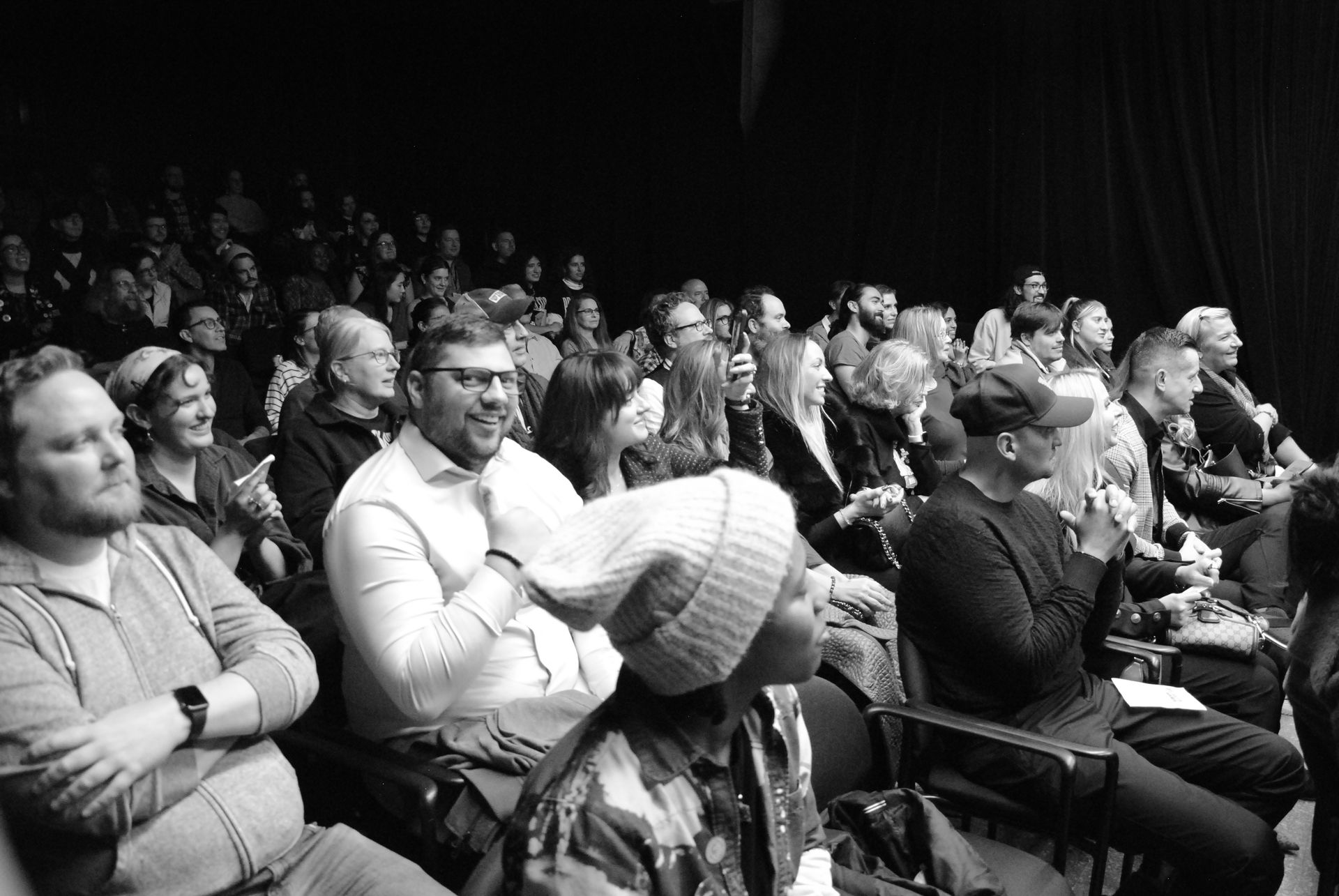 Audience in a dark theater clapping. Many faces and expressions, mostly focused on the stage.