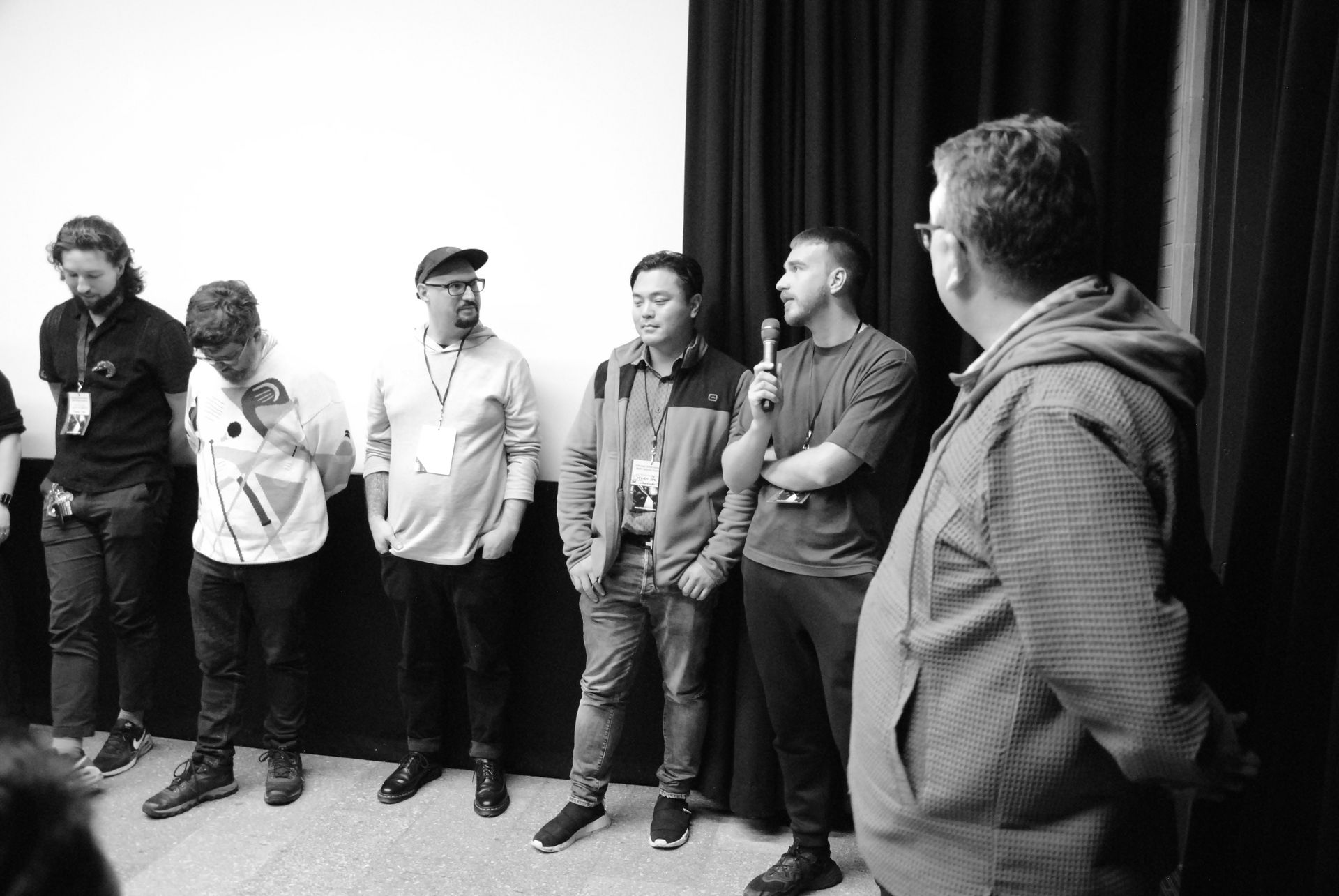Six men on stage, one with a microphone. Some have lanyards. Black and white photo.