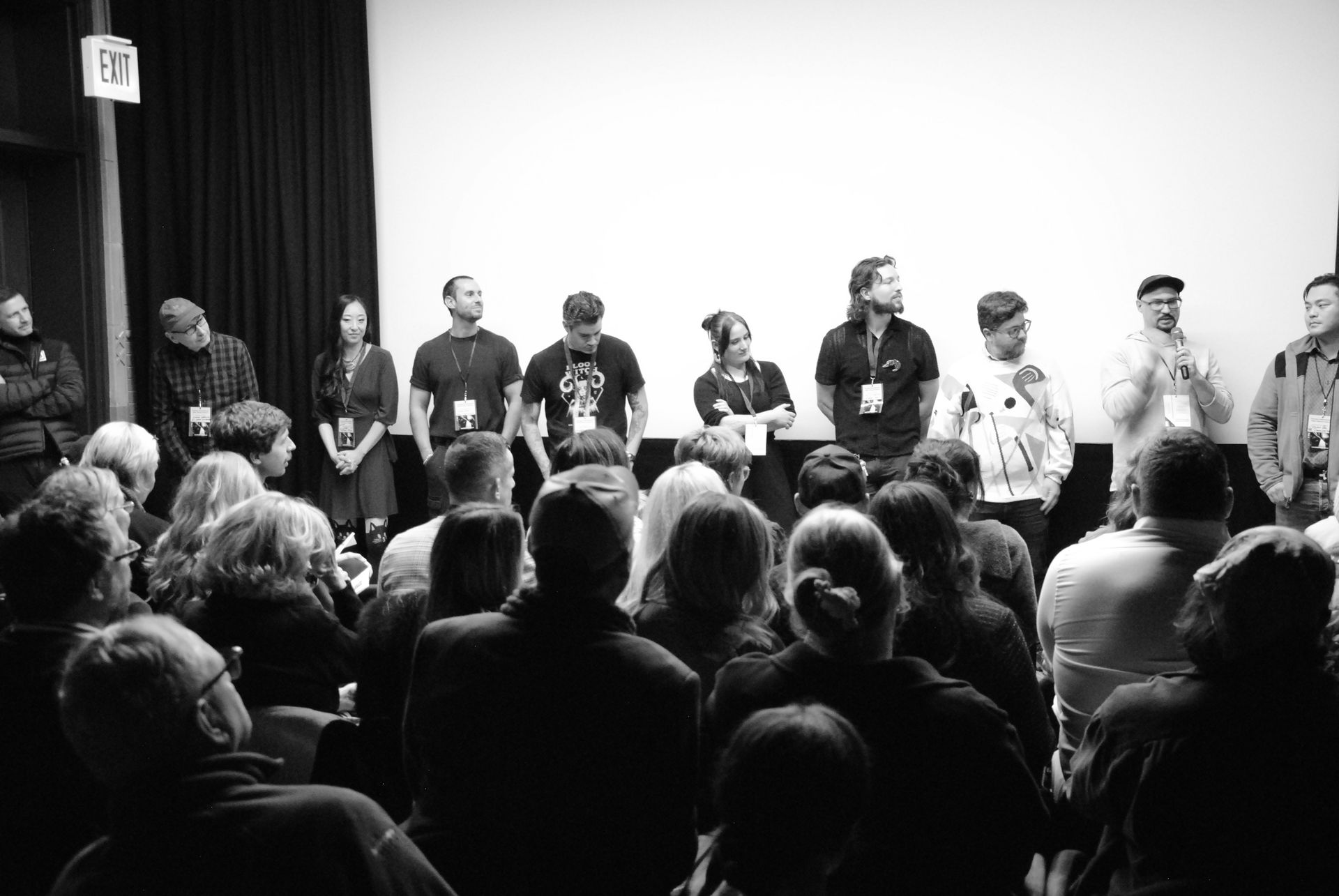 Panel of people standing before a seated audience, facing a screen. Black and white.