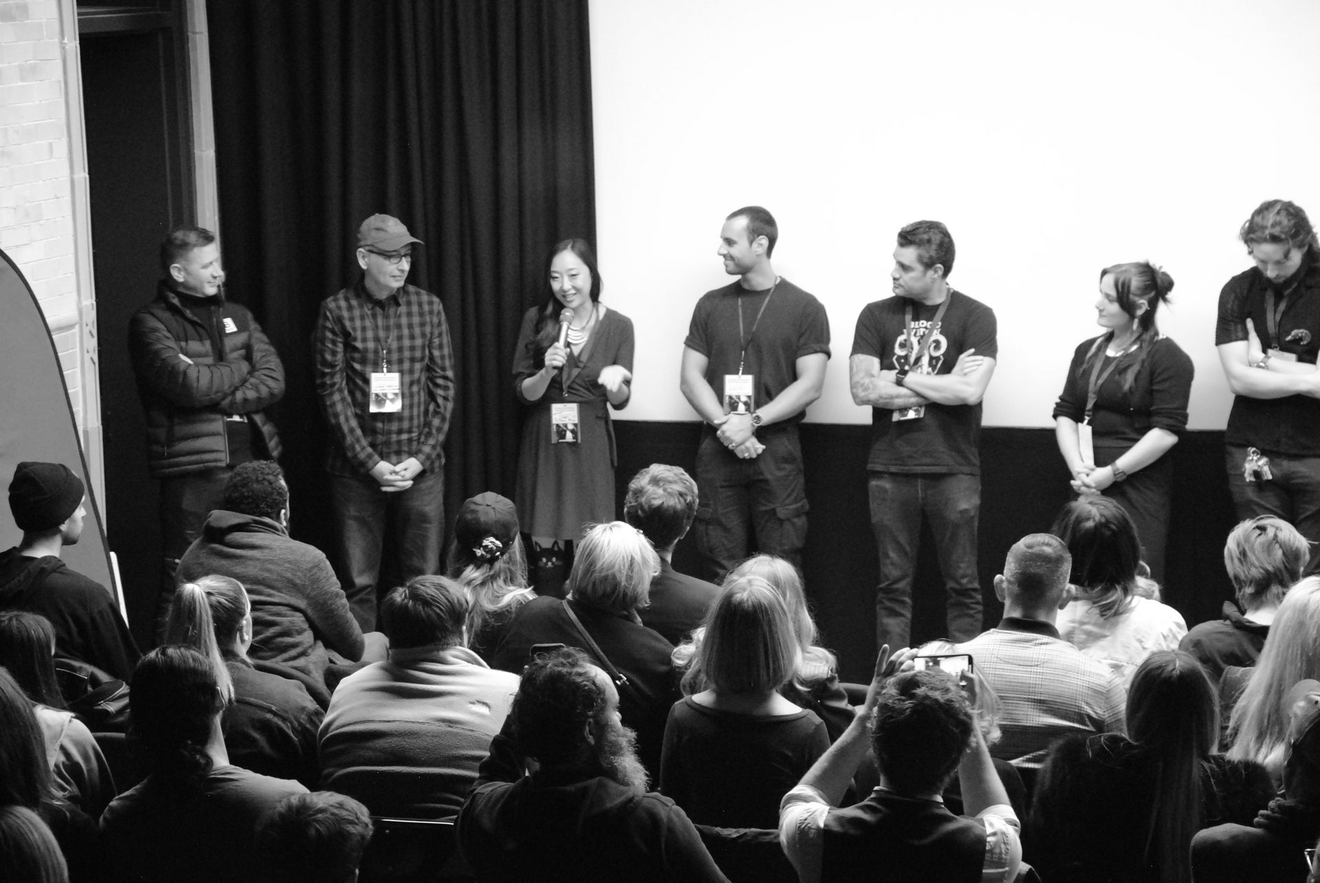 Panel of seven people on a stage in front of a crowd, microphones, white screen.