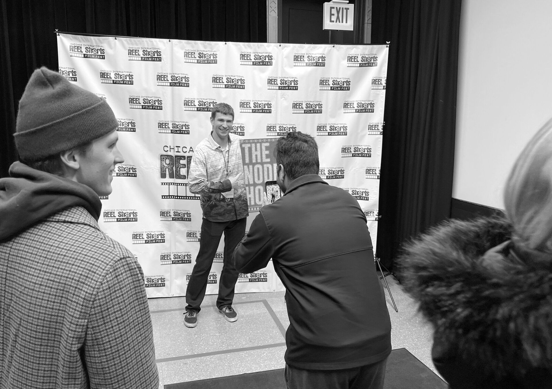 People at event, taking photo of man in front of backdrop; man smiling.