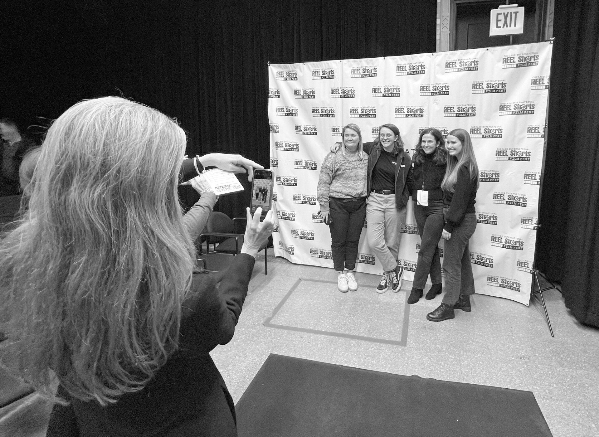 Woman taking photo of four smiling women in front of a branded backdrop.