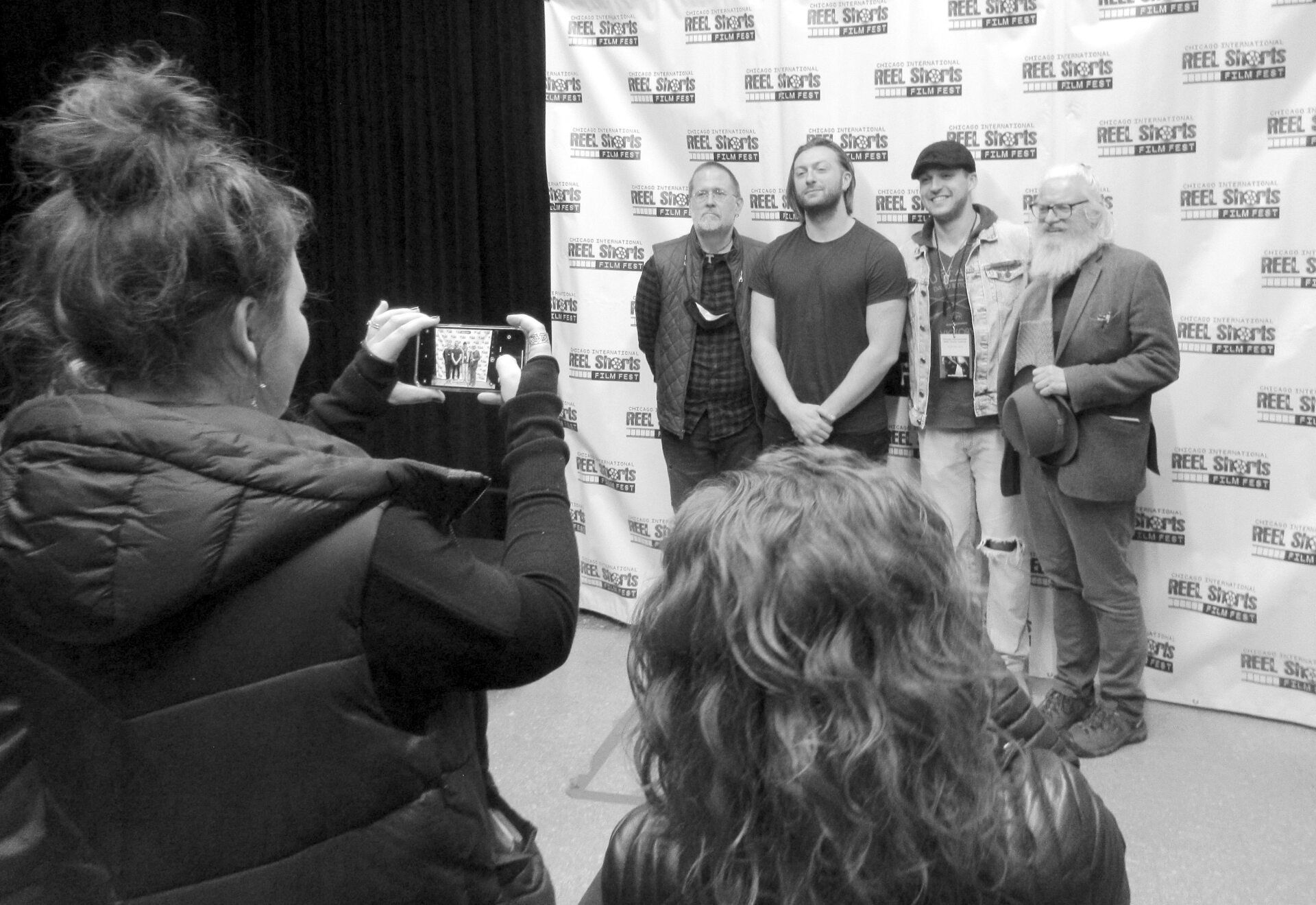 Woman taking photo of four people standing in front of a backdrop. Black and white.