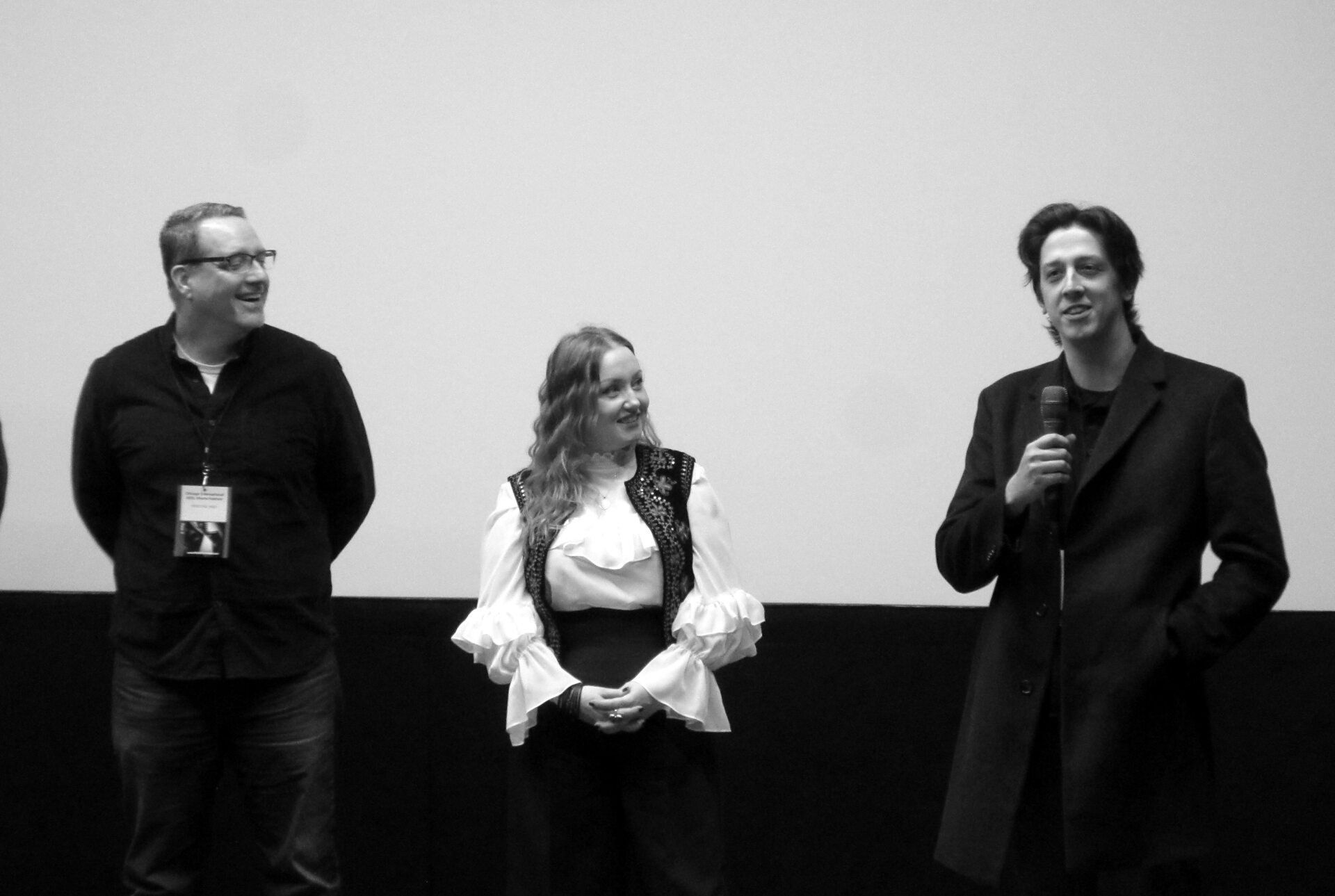Three people on stage: man with glasses, woman in vest, man with microphone, all smiling.