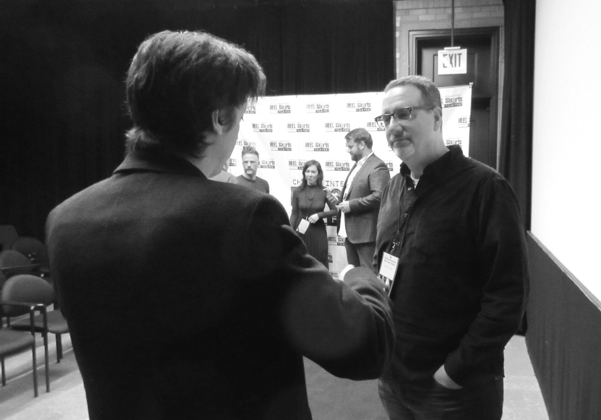 Two men shaking hands inside a venue; group of people in background. Black and white photo.