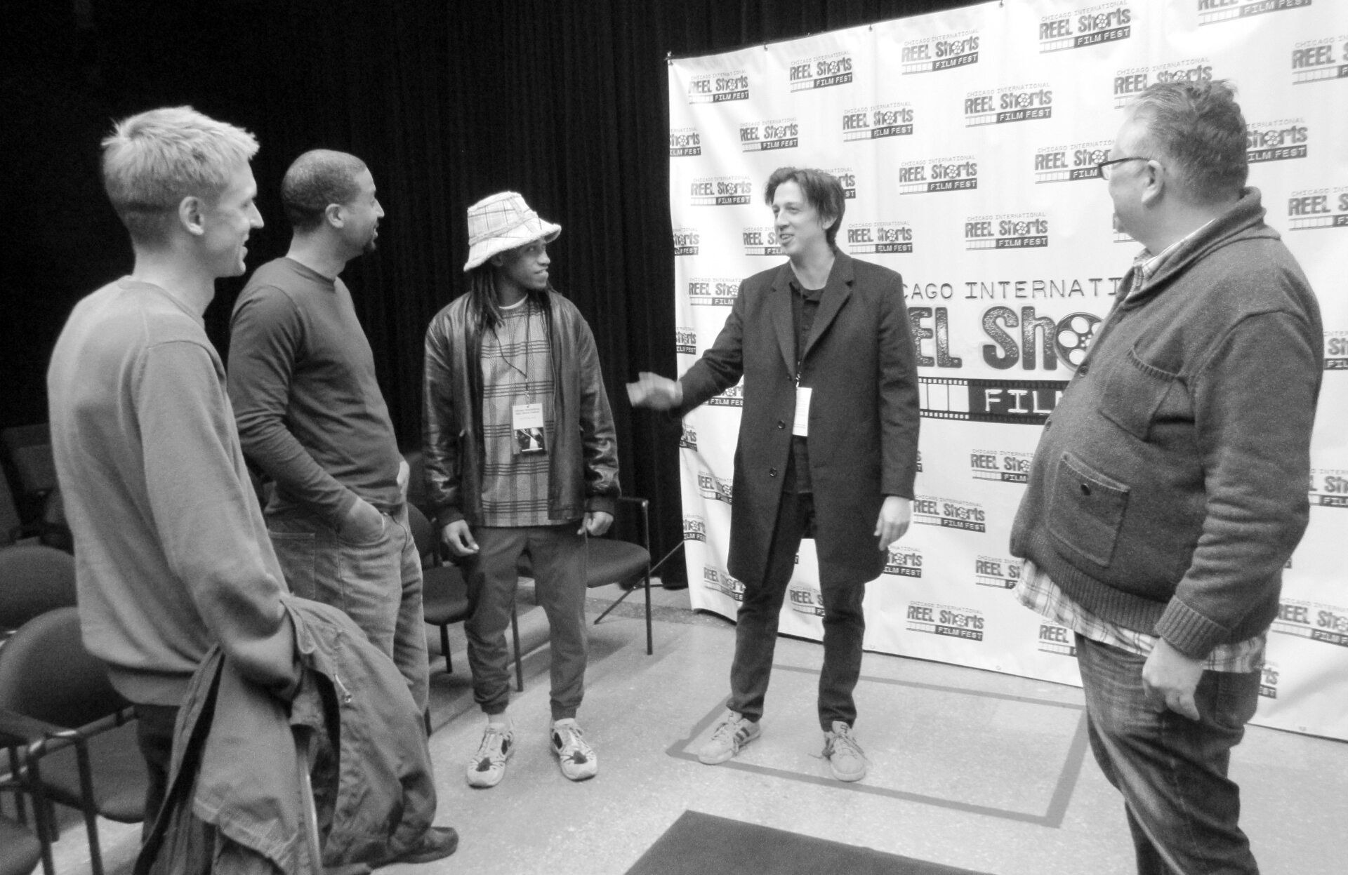 Five people stand in a room, one in a hat gestures. Event banner in the background.