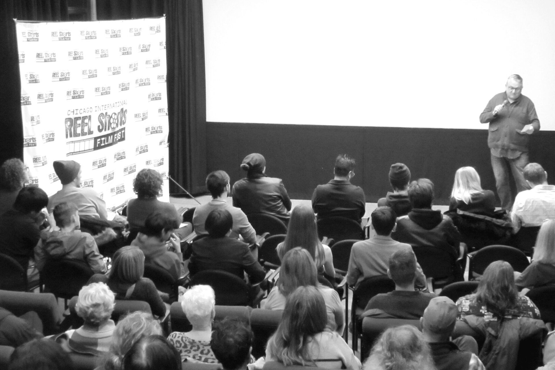 Audience watching a speaker in a theater. A banner on the left reads 