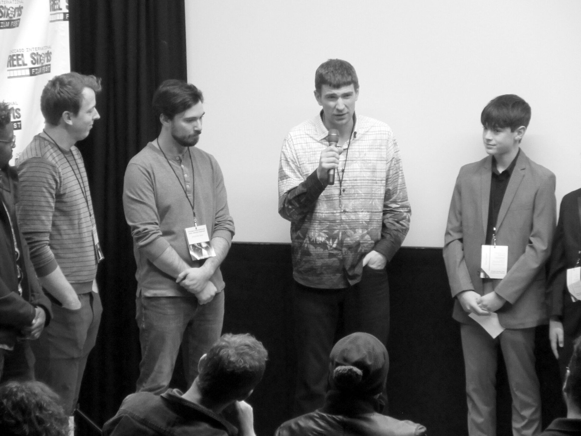 Group of men on a stage, one speaking into a microphone, facing an audience, black and white.