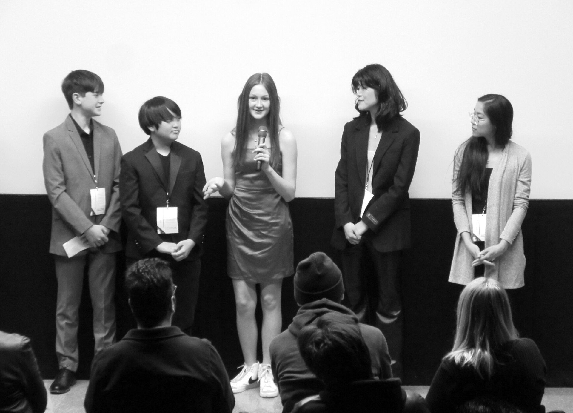 A group of teens on a stage, girl holding a microphone addressing audience. Black and white.
