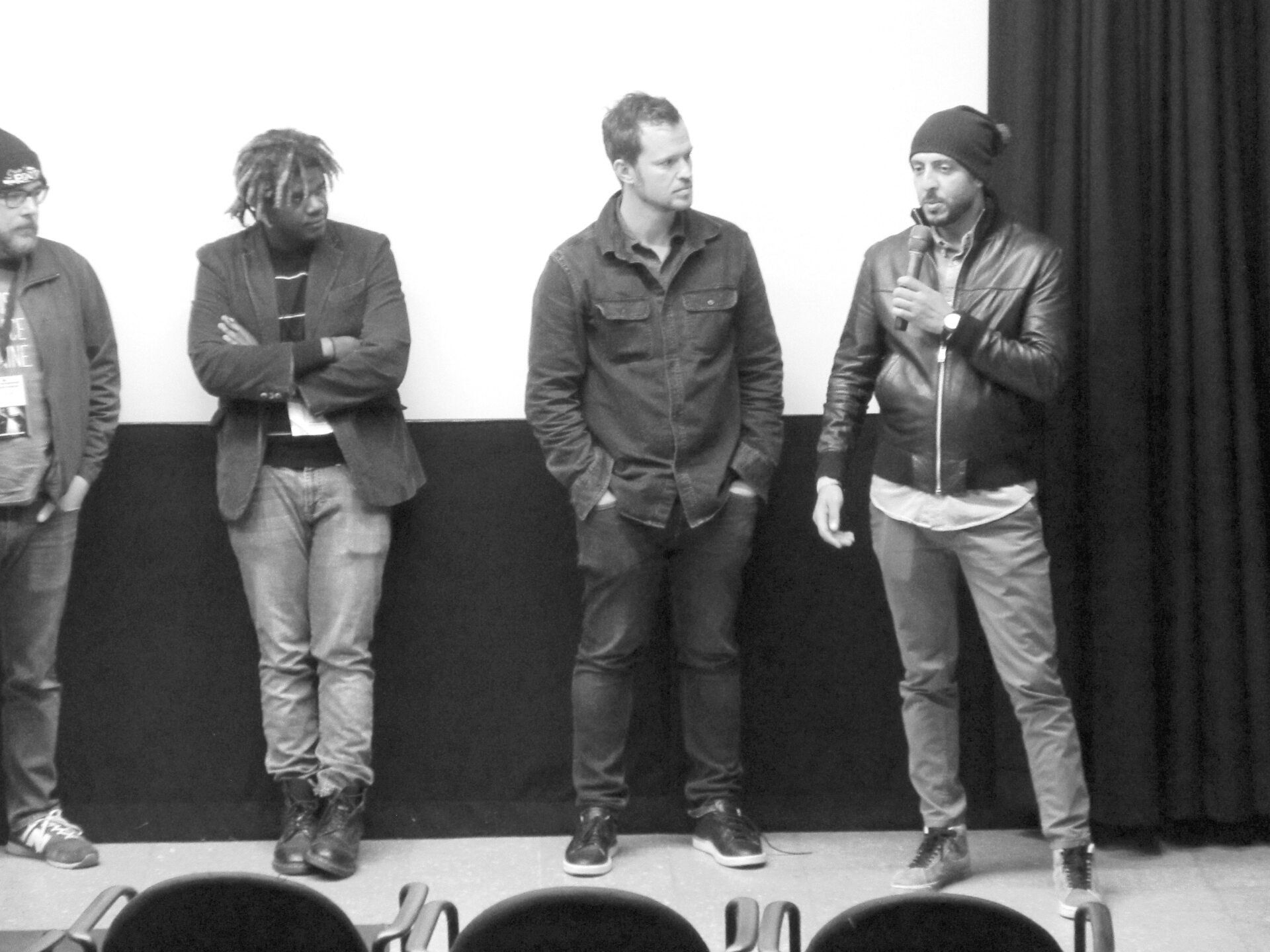 Four people standing in front of a screen, one speaking into a microphone. Black and white.
