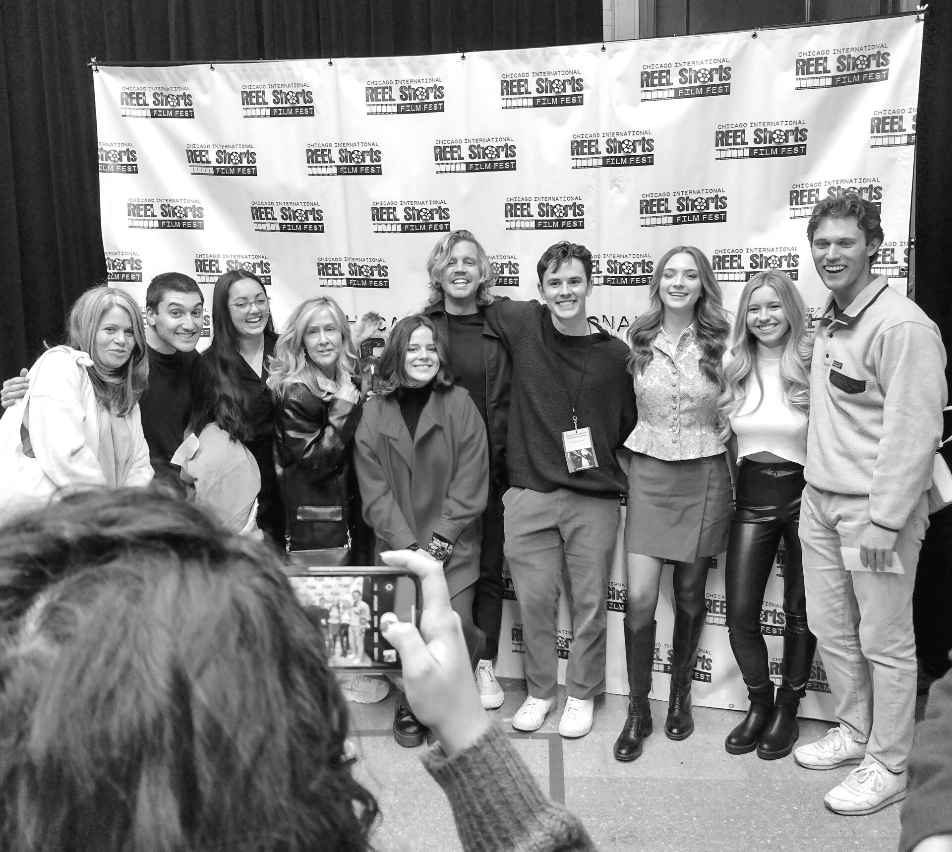Group poses for a photo in front of a banner, smiling. People are holding a phone, inside.