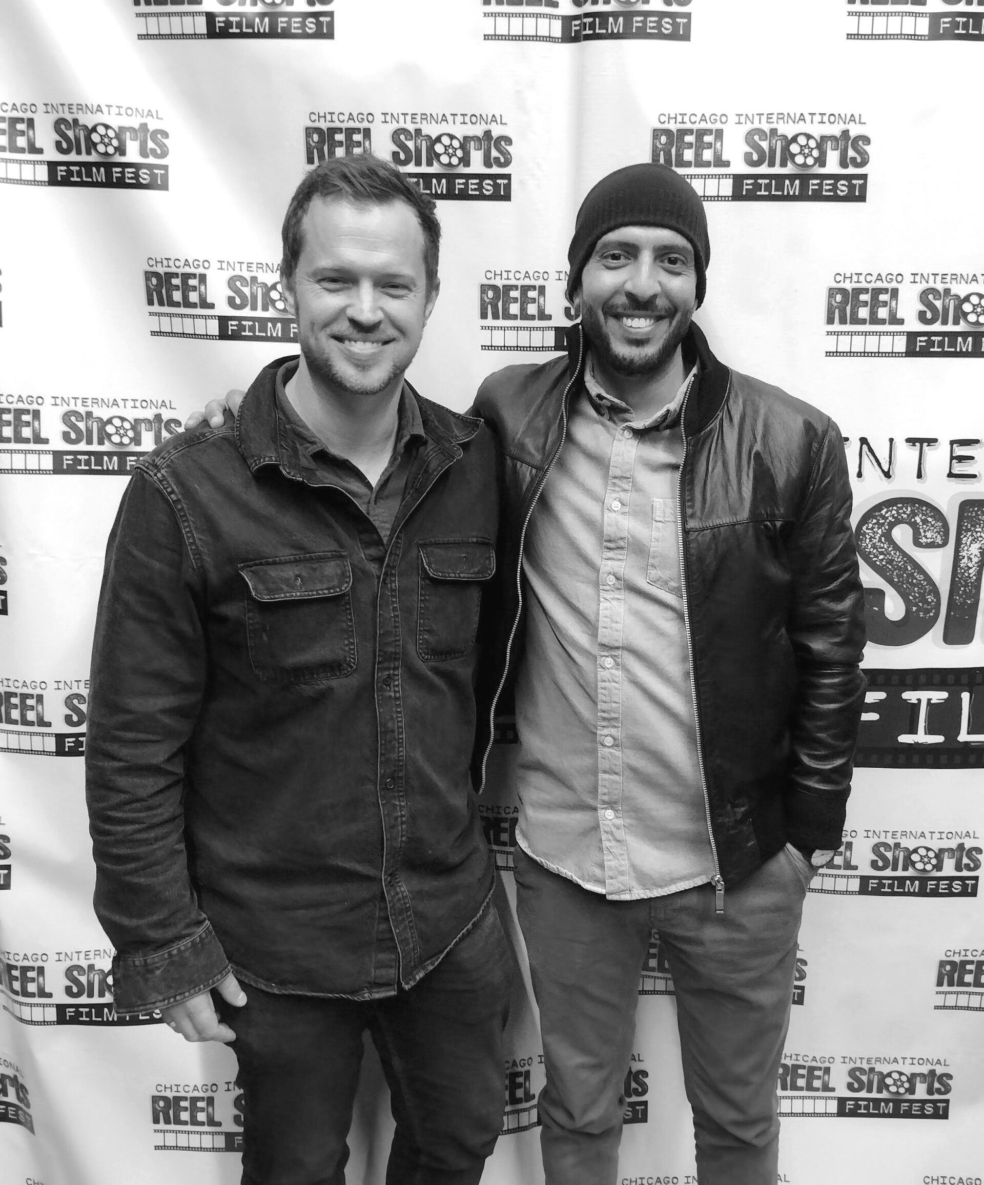 Two men smiling, posed together at a film festival, the background with event banners.