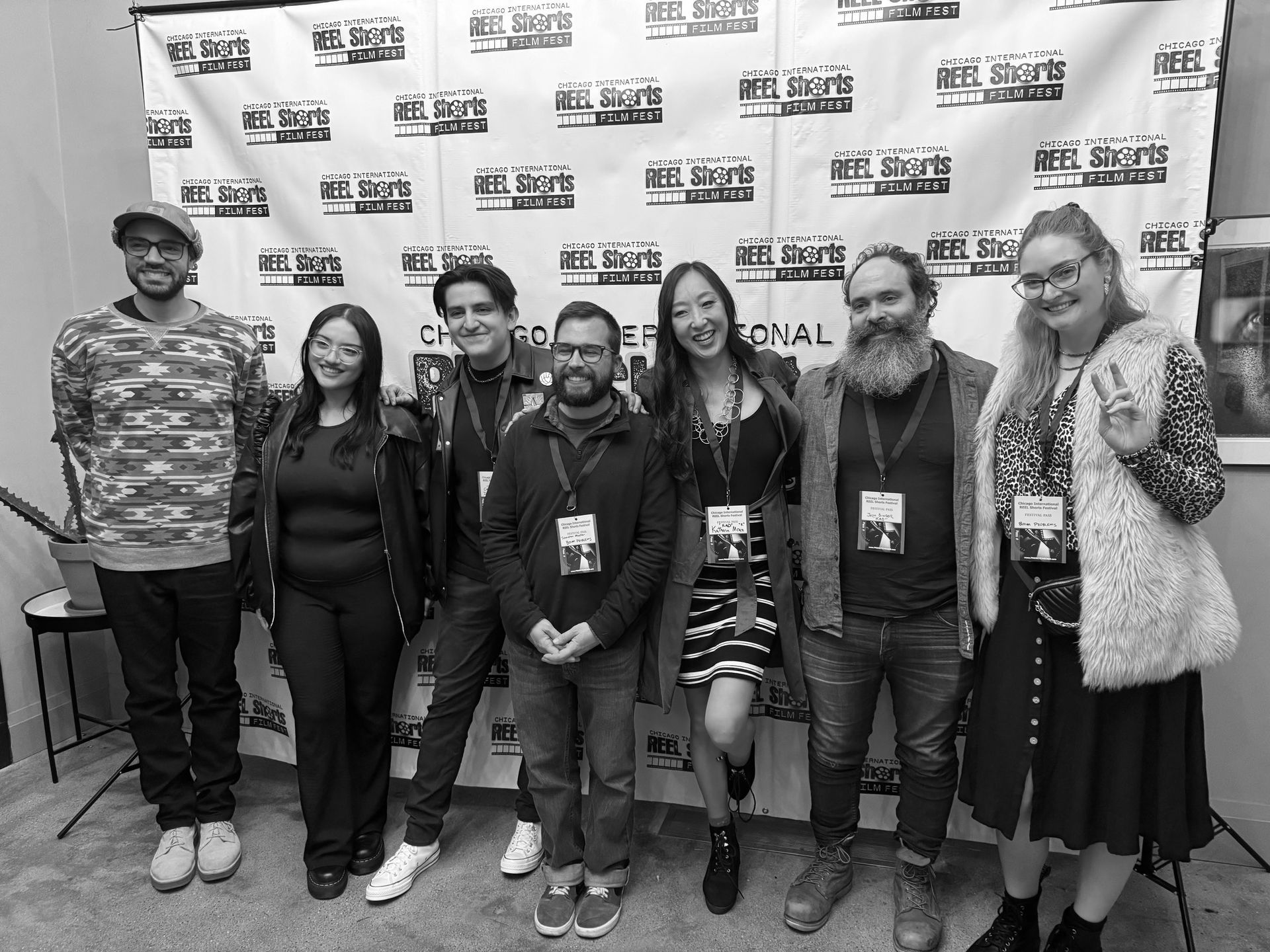 Group poses in front of a backdrop. Seven people, diverse, smiling. Festival event. Black and white.