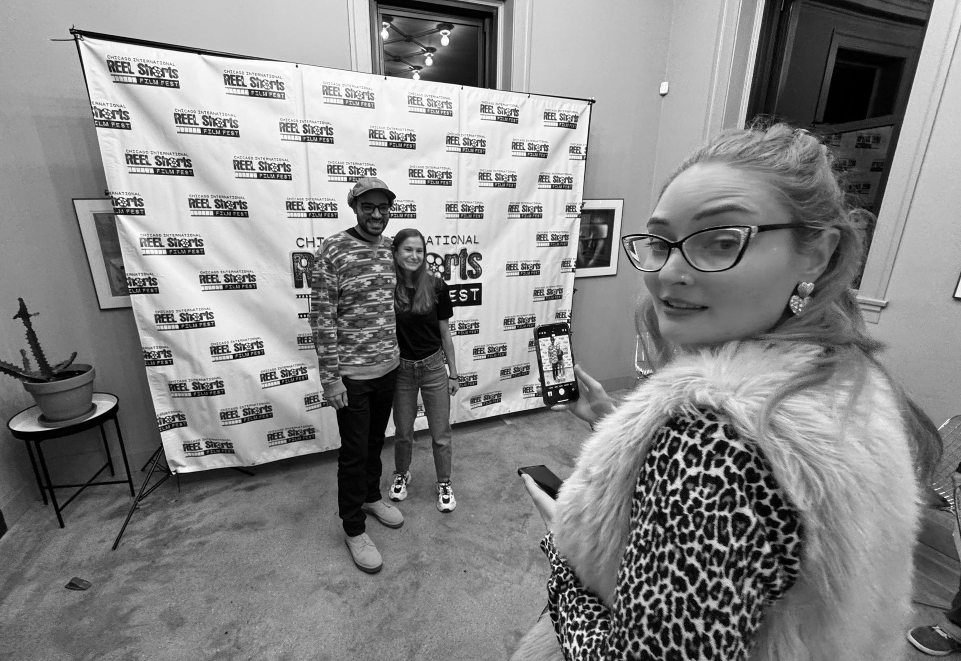 Woman taking photo of couple in front of a backdrop at an event. Black and white.