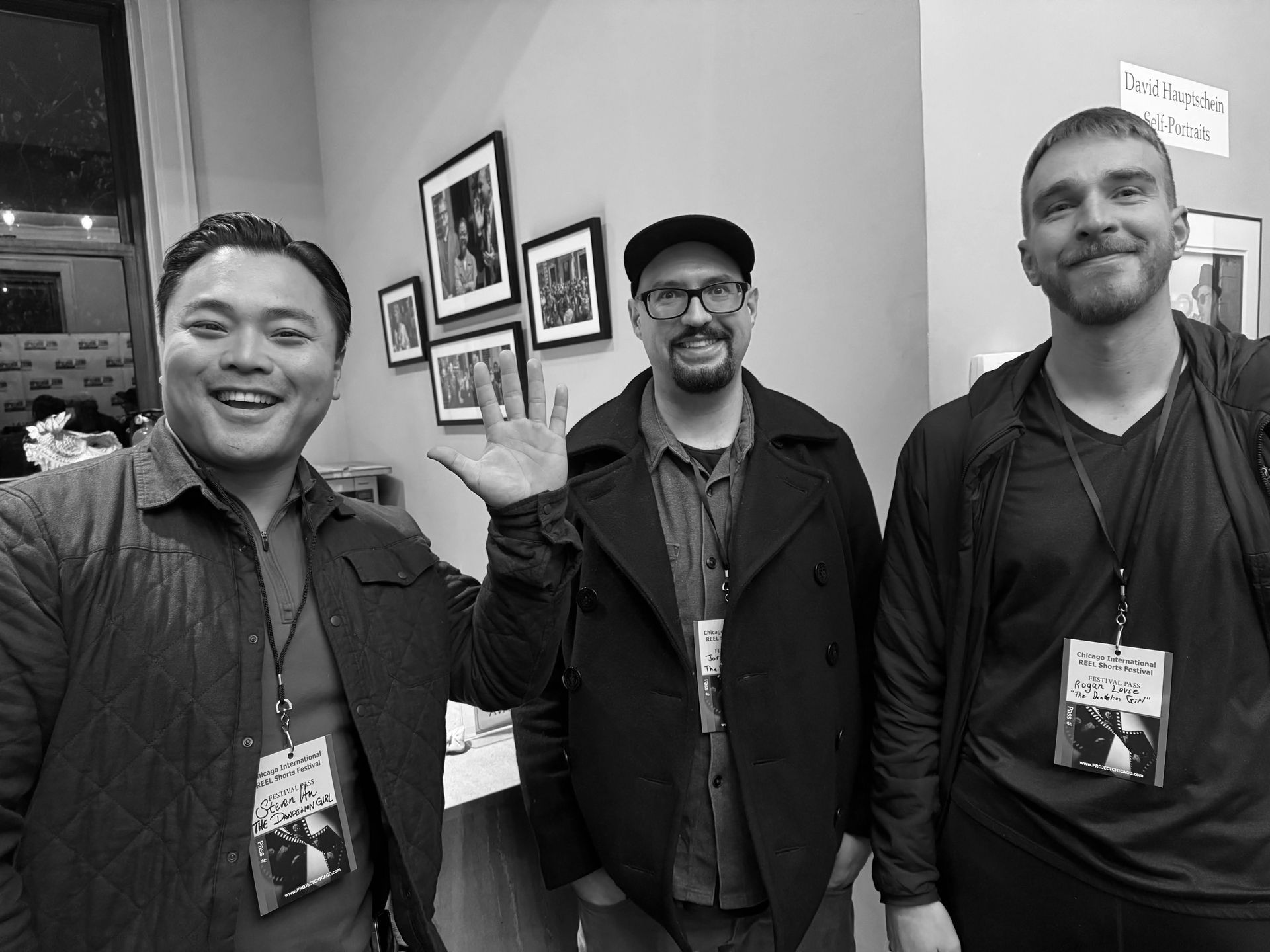 Three men posing indoors, one waving. They wear lanyards, near framed photos, with smiles.