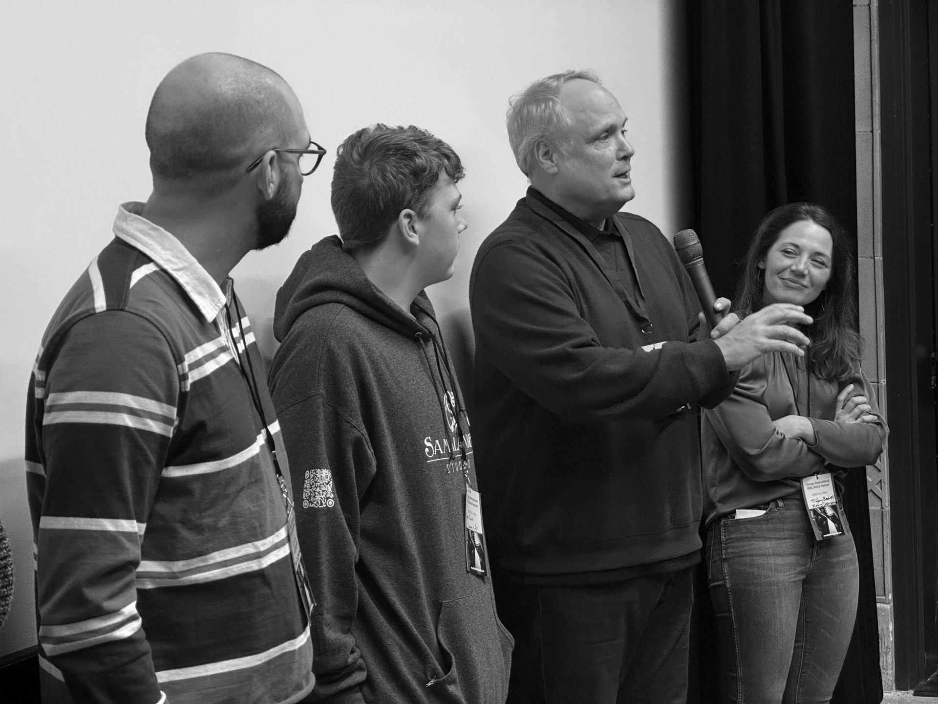 Four people stand near a doorway, one speaking into a microphone. Black and white photo.