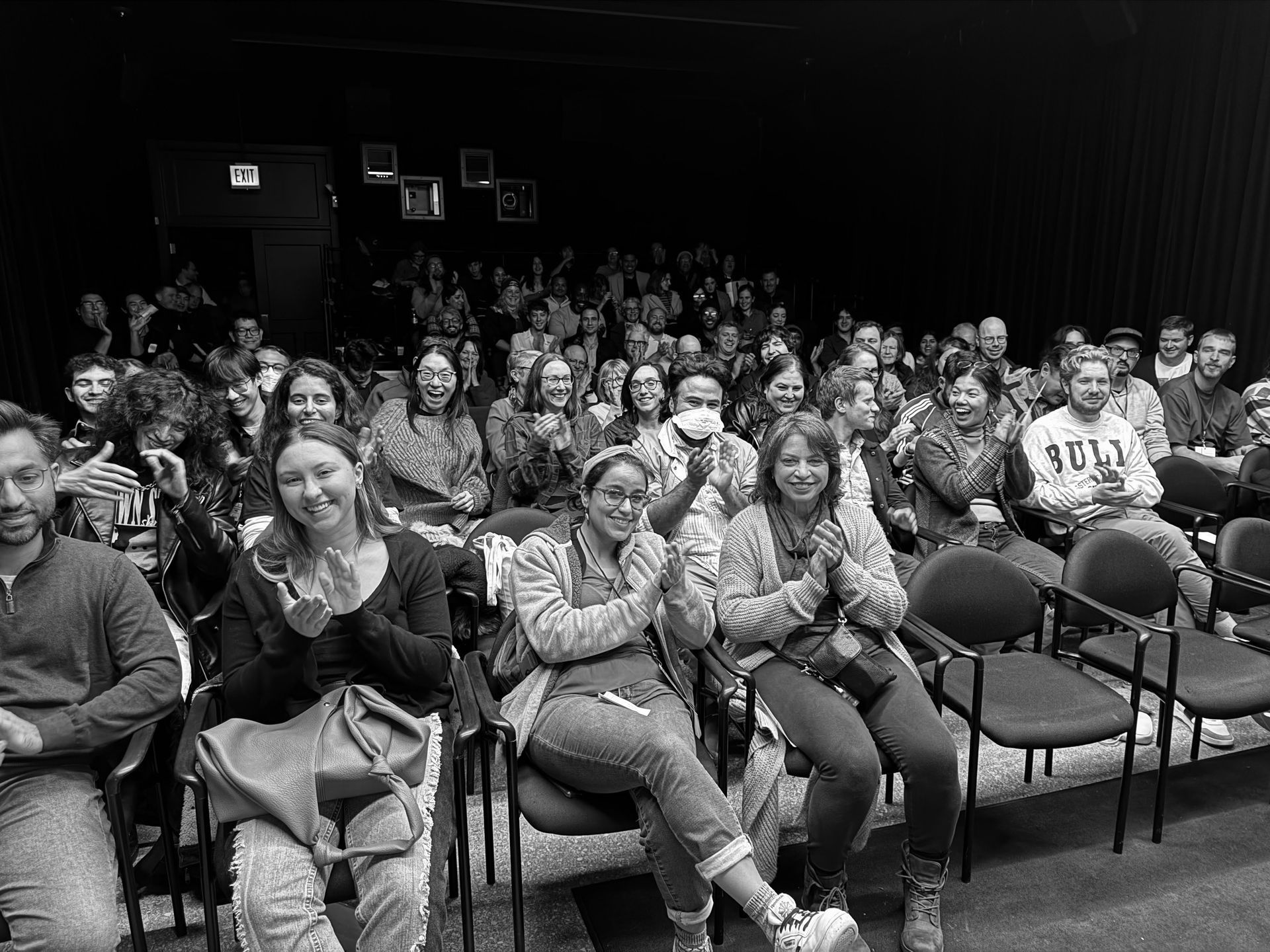 Audience in a dark auditorium clapping. Several people in front row are smiling and applauding.