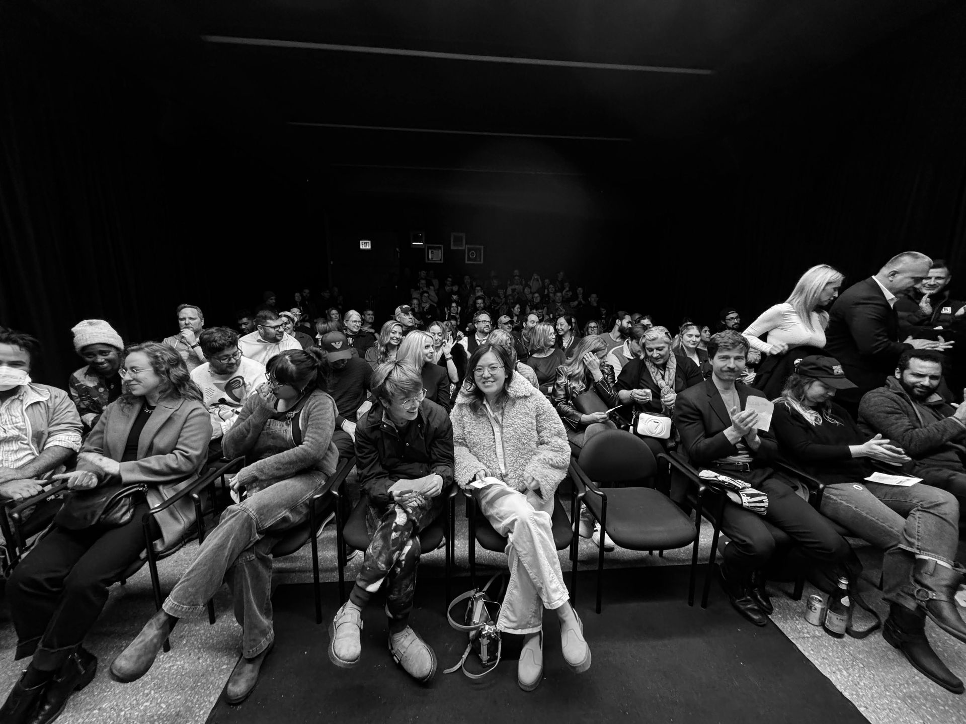 Audience seated in a dimly lit theater. People of diverse ages and dress observe something from their seats.