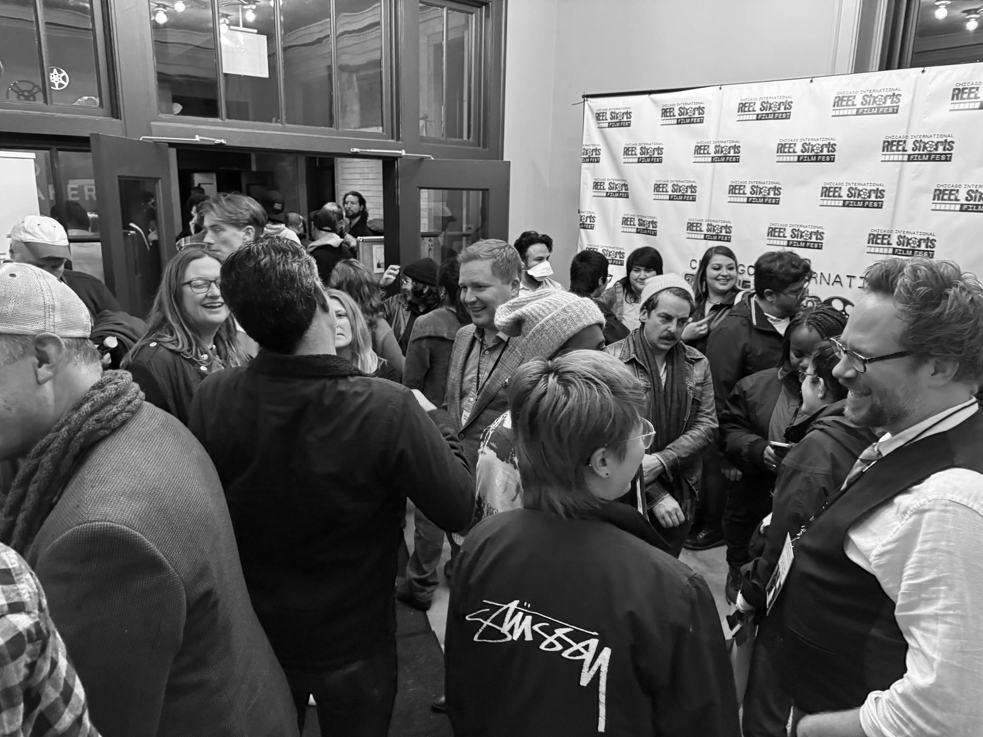 Crowd of people in a doorway, likely at an event. Many smiling, some wearing coats. Black and white.