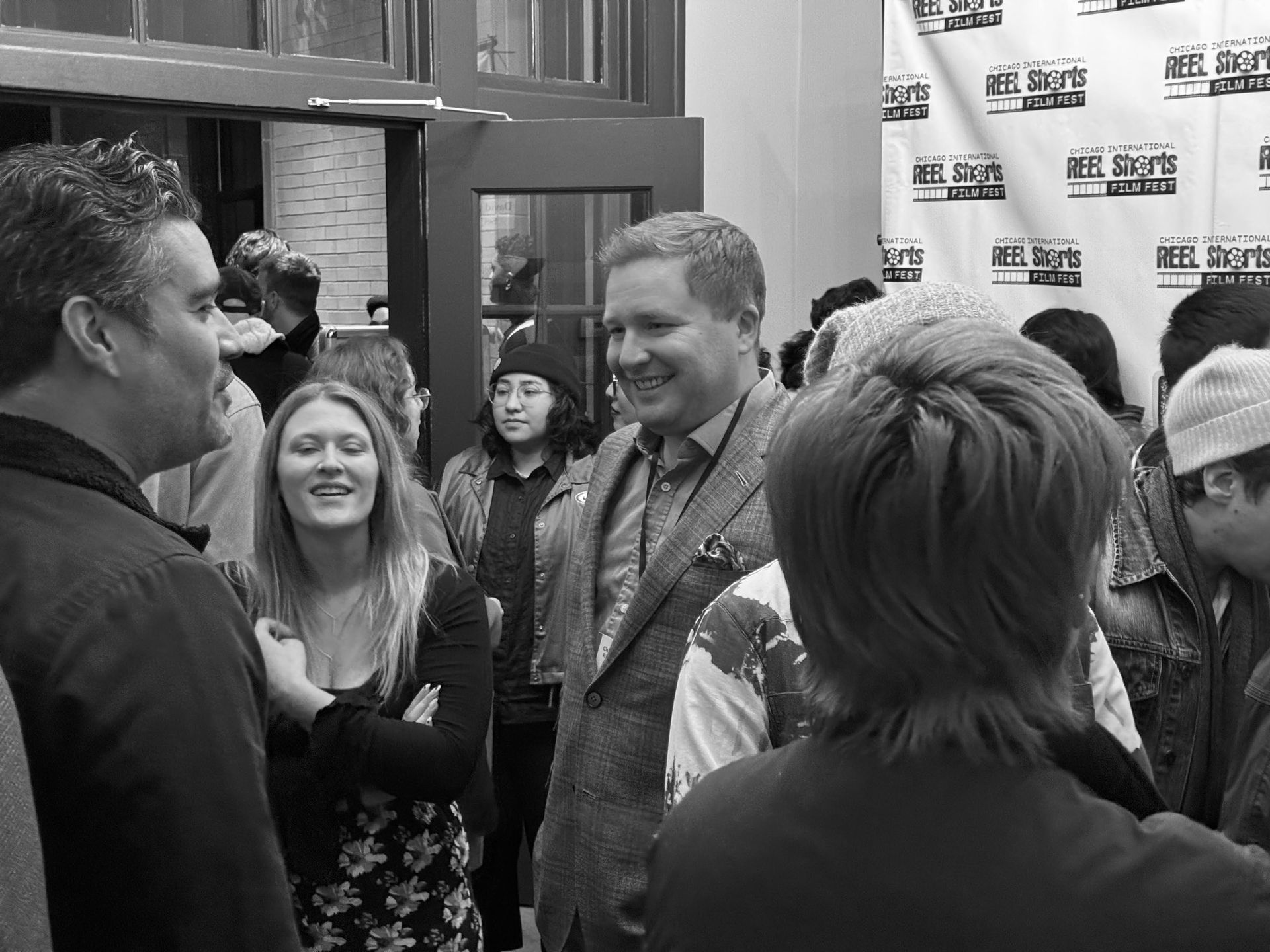 People gathered inside, smiling and talking, likely at an event. Black and white photo.