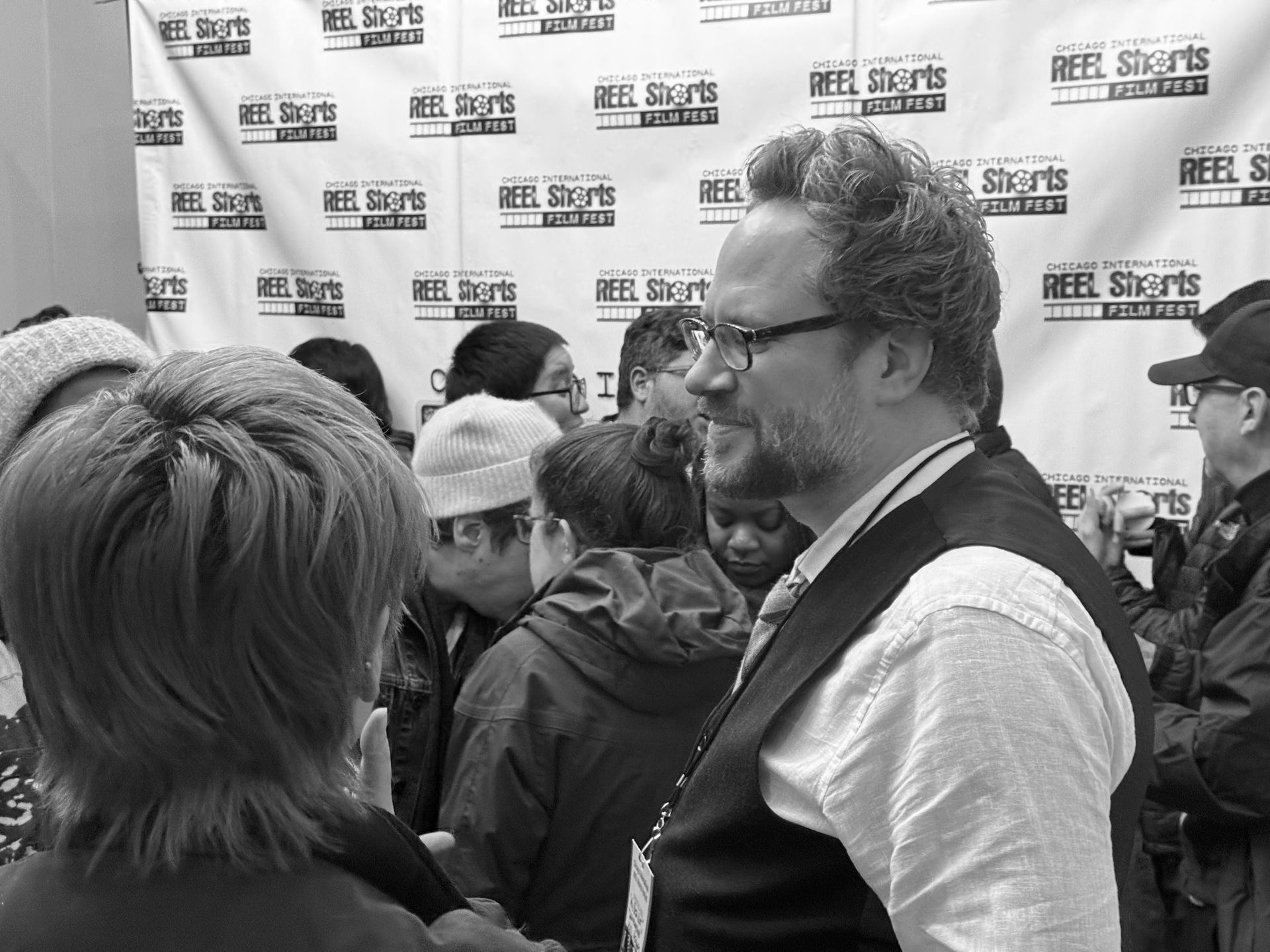 Man in vest speaks to someone; people and backdrop with logos.