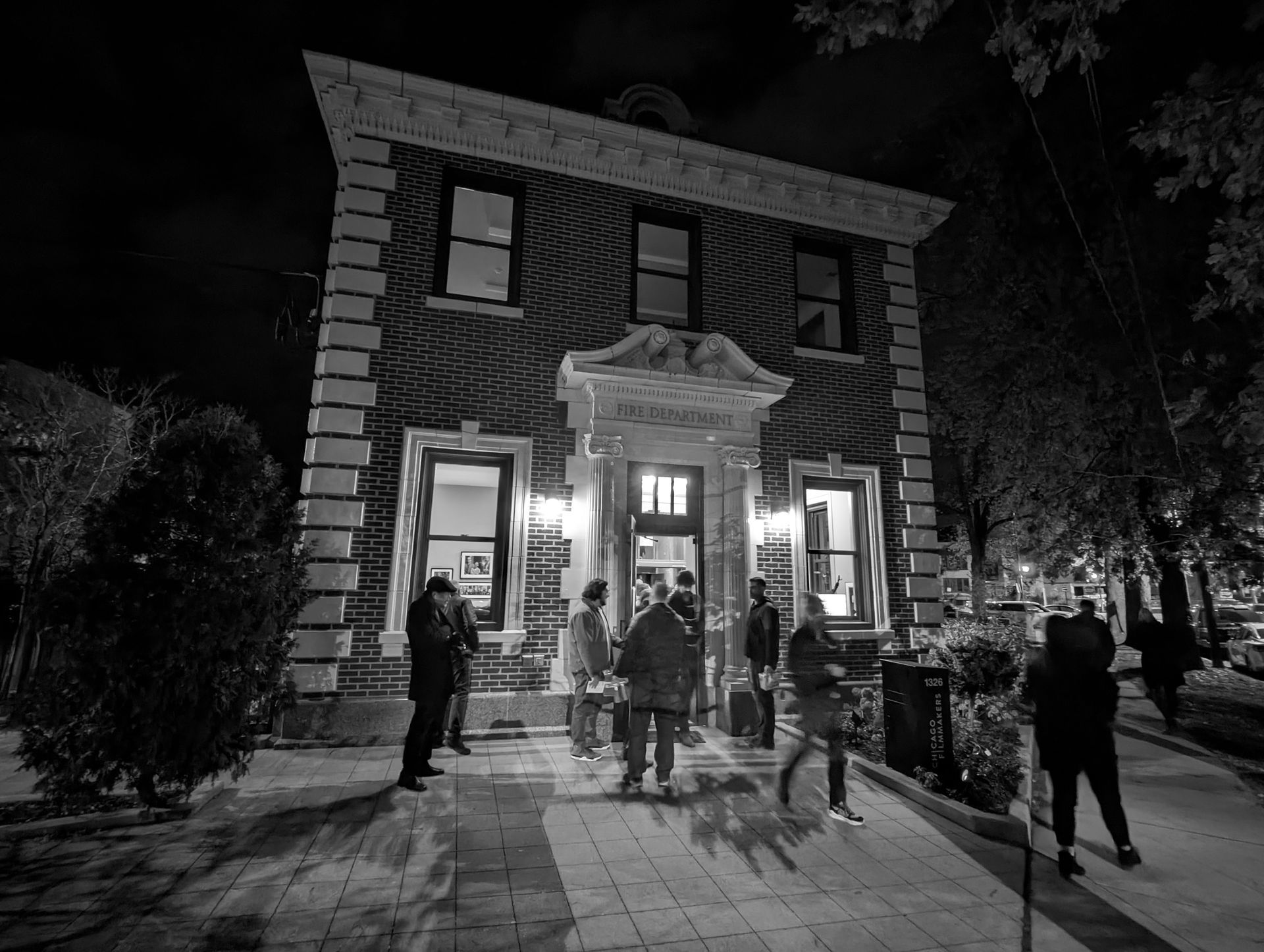 A two-story brick building at night, with people gathered outside; lit windows and doorway.