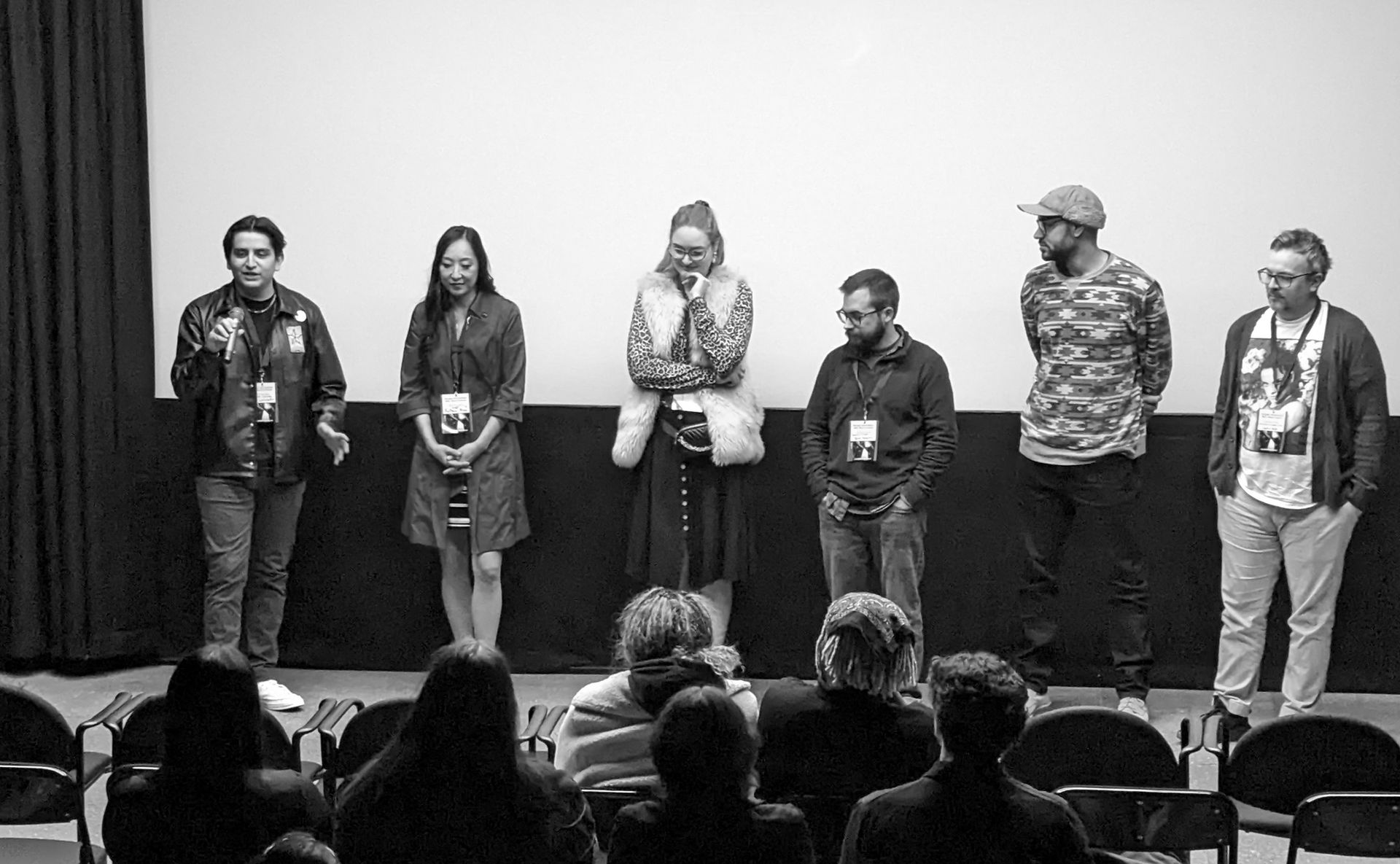 Panel of seven people on stage at a theater, facing the audience. Black and white.