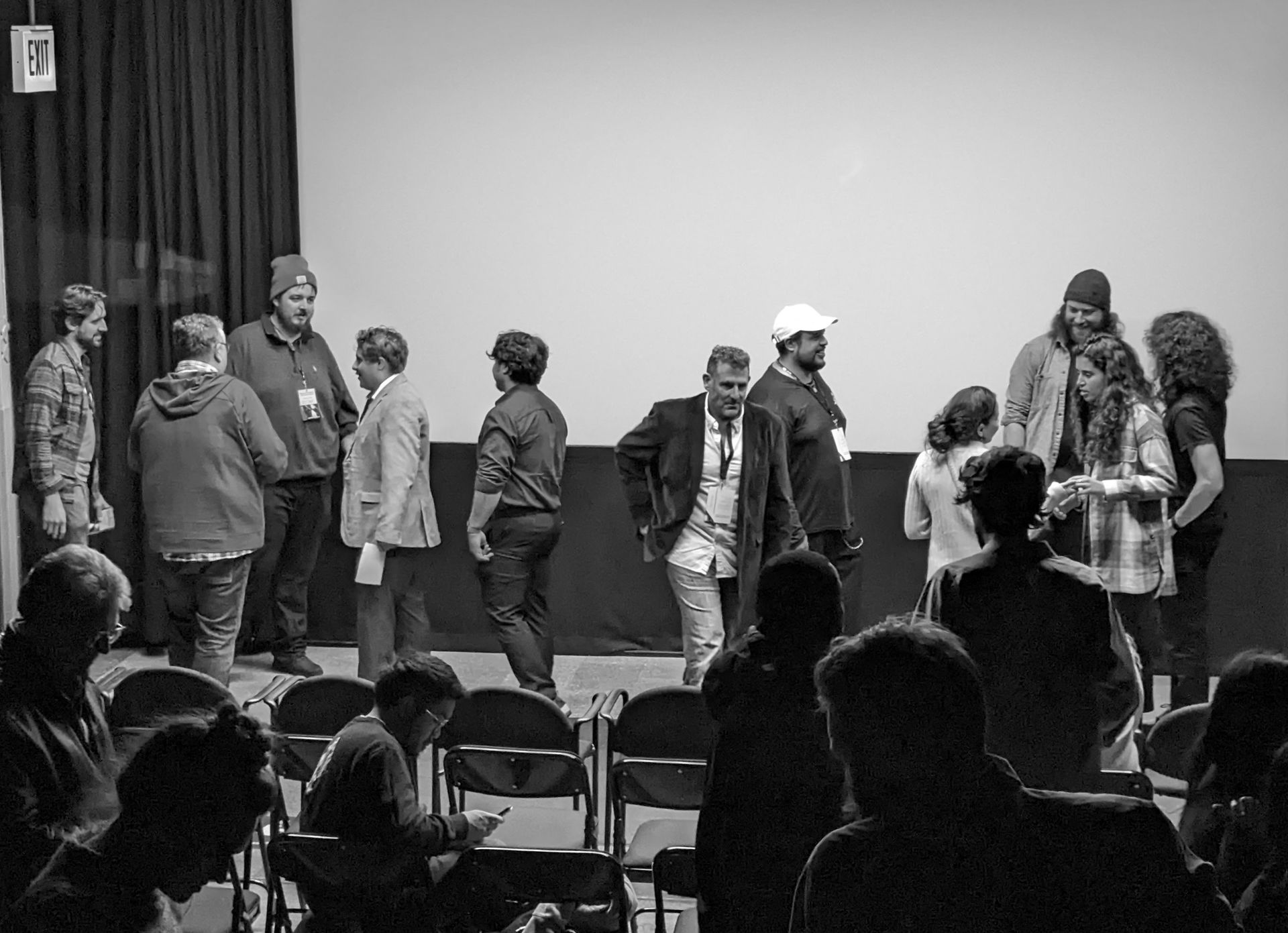 A group of people on stage in front of a white screen; audience members in chairs in foreground.