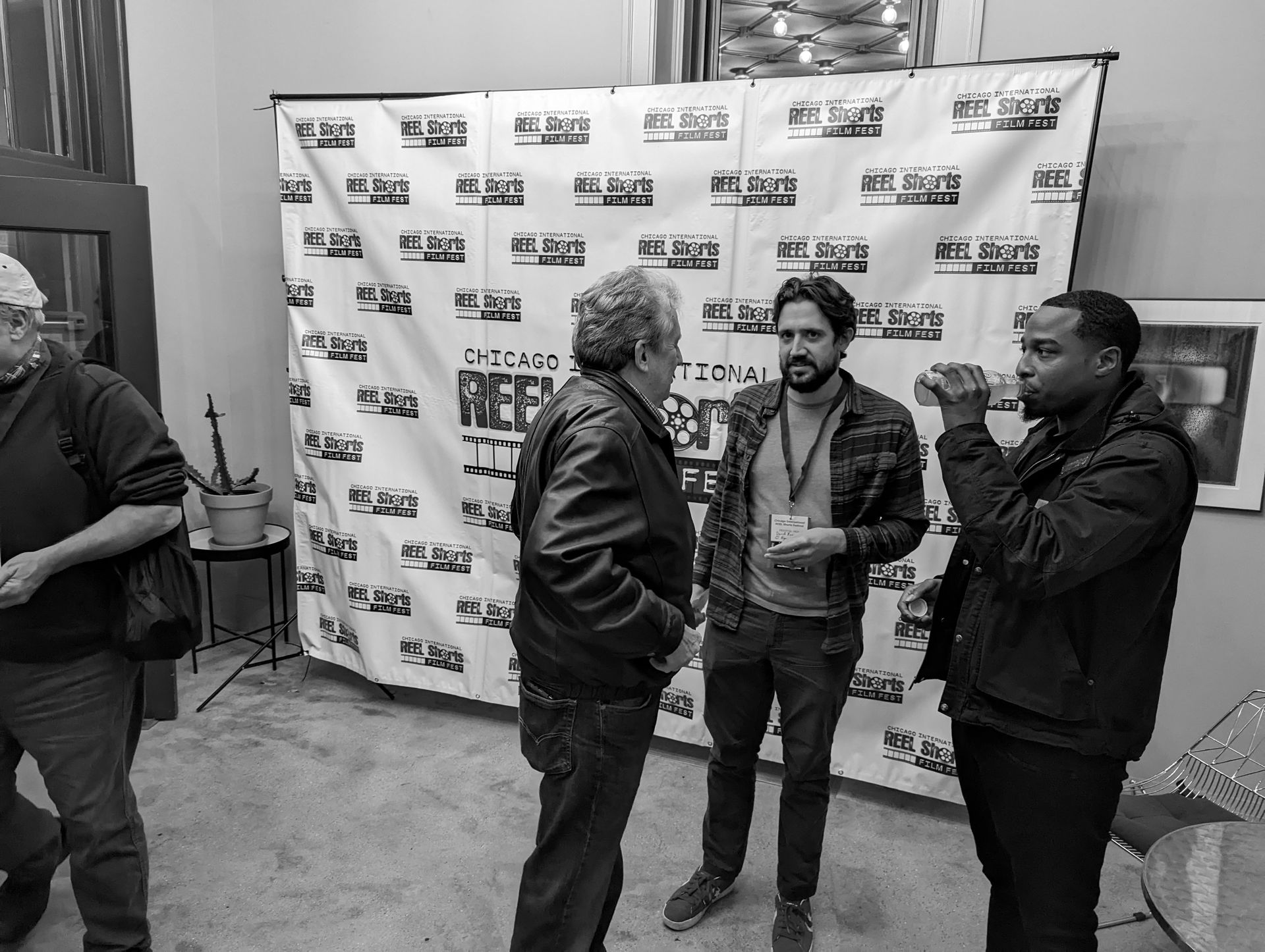 Four men stand in a room with a backdrop. Two men converse, the third holds a drink, and the fourth walks by.
