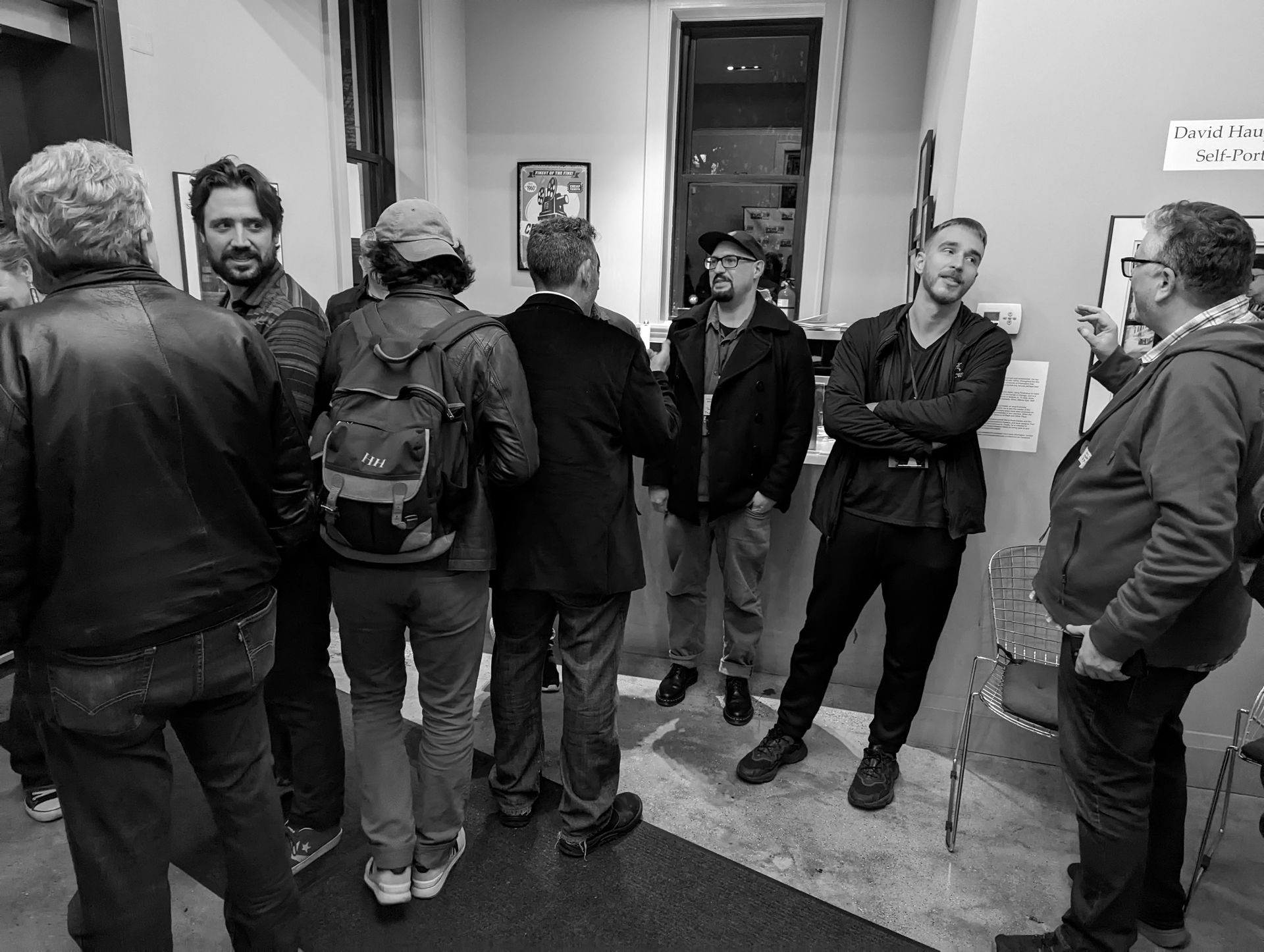 Group of people socializing inside an art gallery. Some are looking at artwork, others are talking. Black and white.