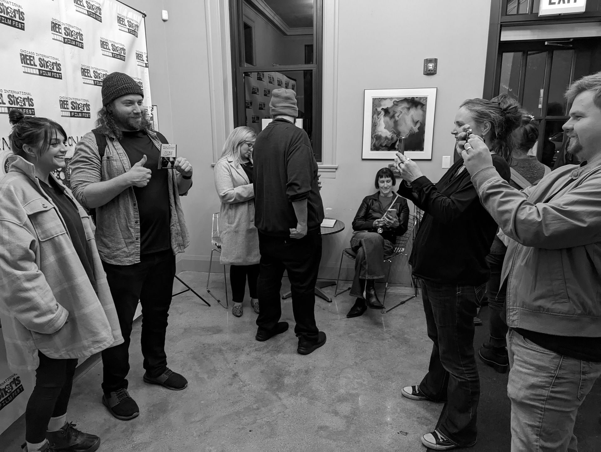 People at an art gallery. Some taking photos, others conversing. Black and white.