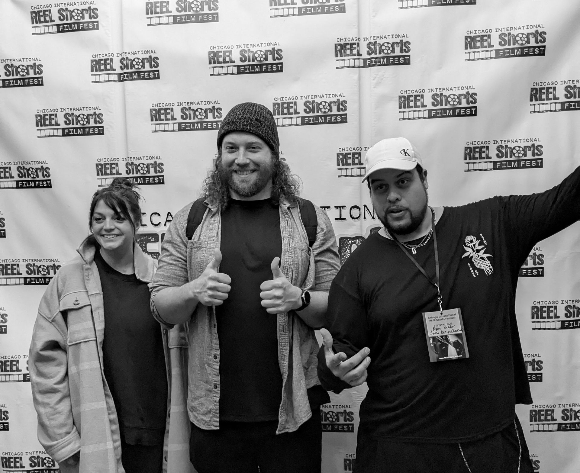 Three people posing with a backdrop. Two men give thumbs up, while a woman stands on the left. The backdrop has the 