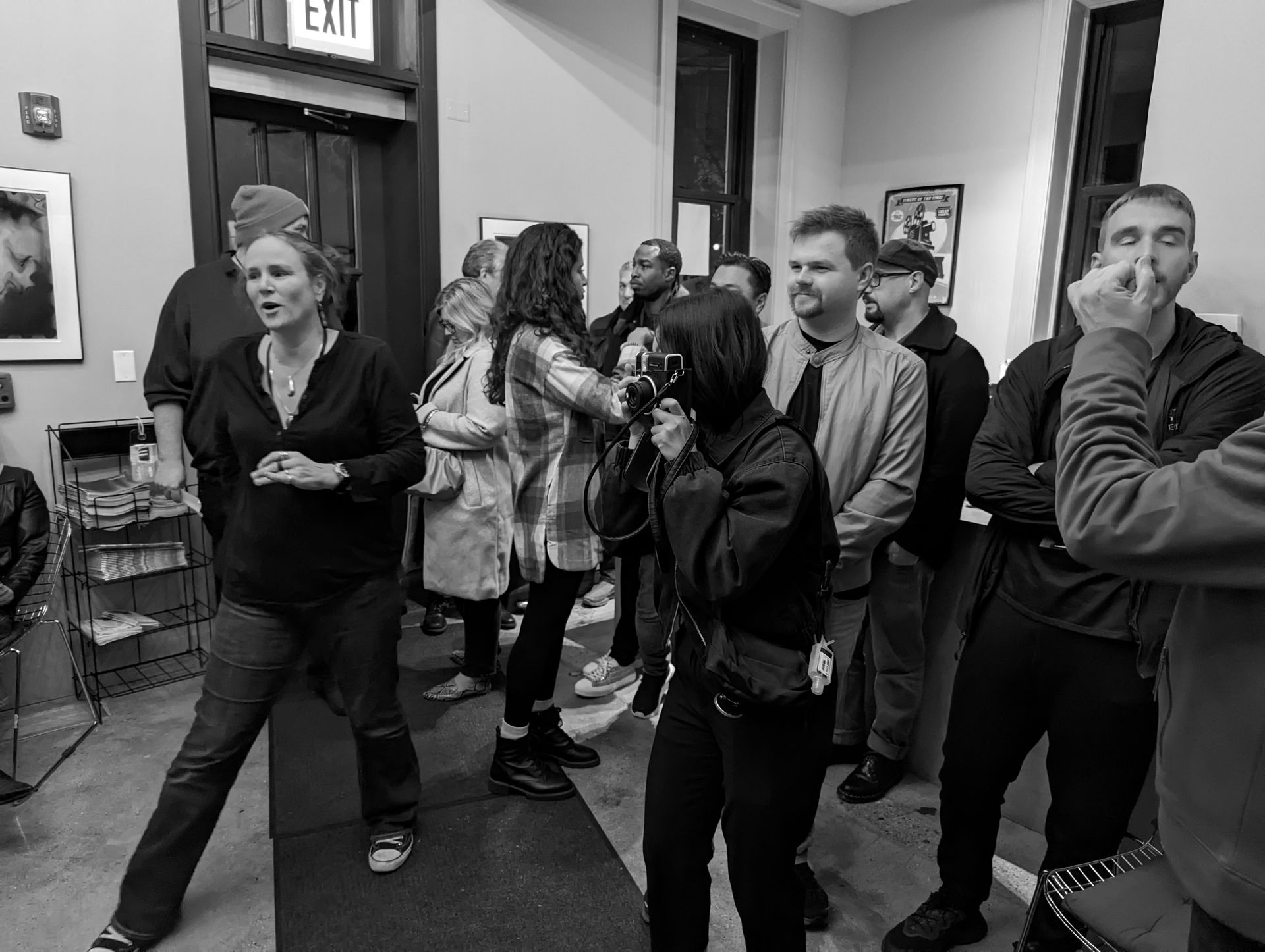 Group of people at an event. Woman in foreground is speaking, another takes photos. Gallery setting, black and white.