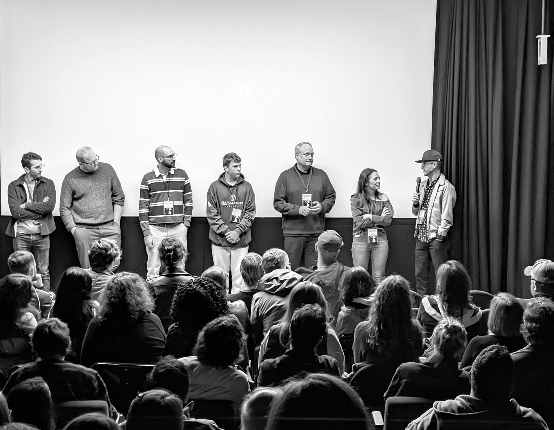 A group of people on a stage in front of an audience. Some hold trophies. Black and white.