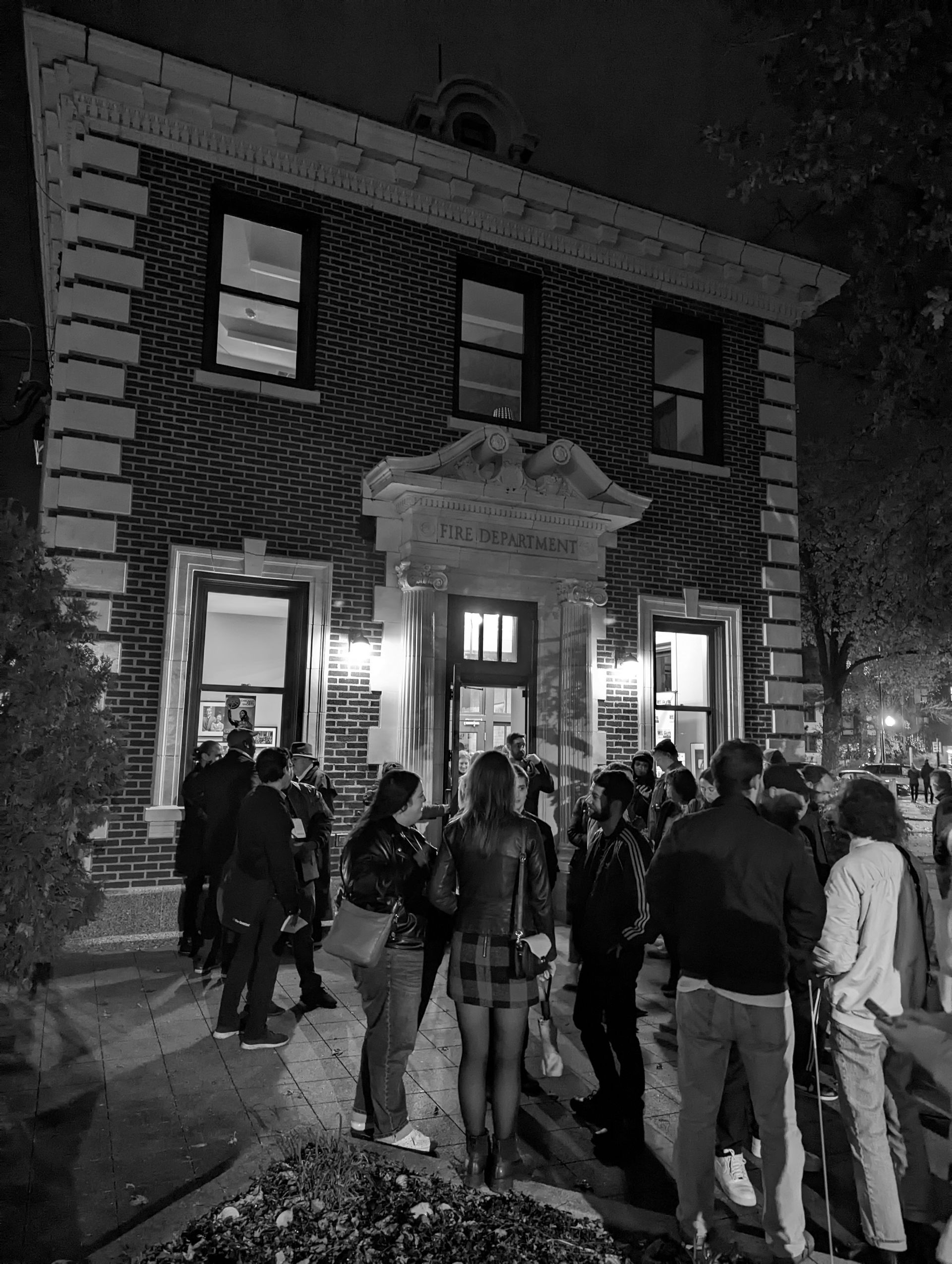 People outside a brick building at night. Lit doorway and windows above. Group of people gathered.