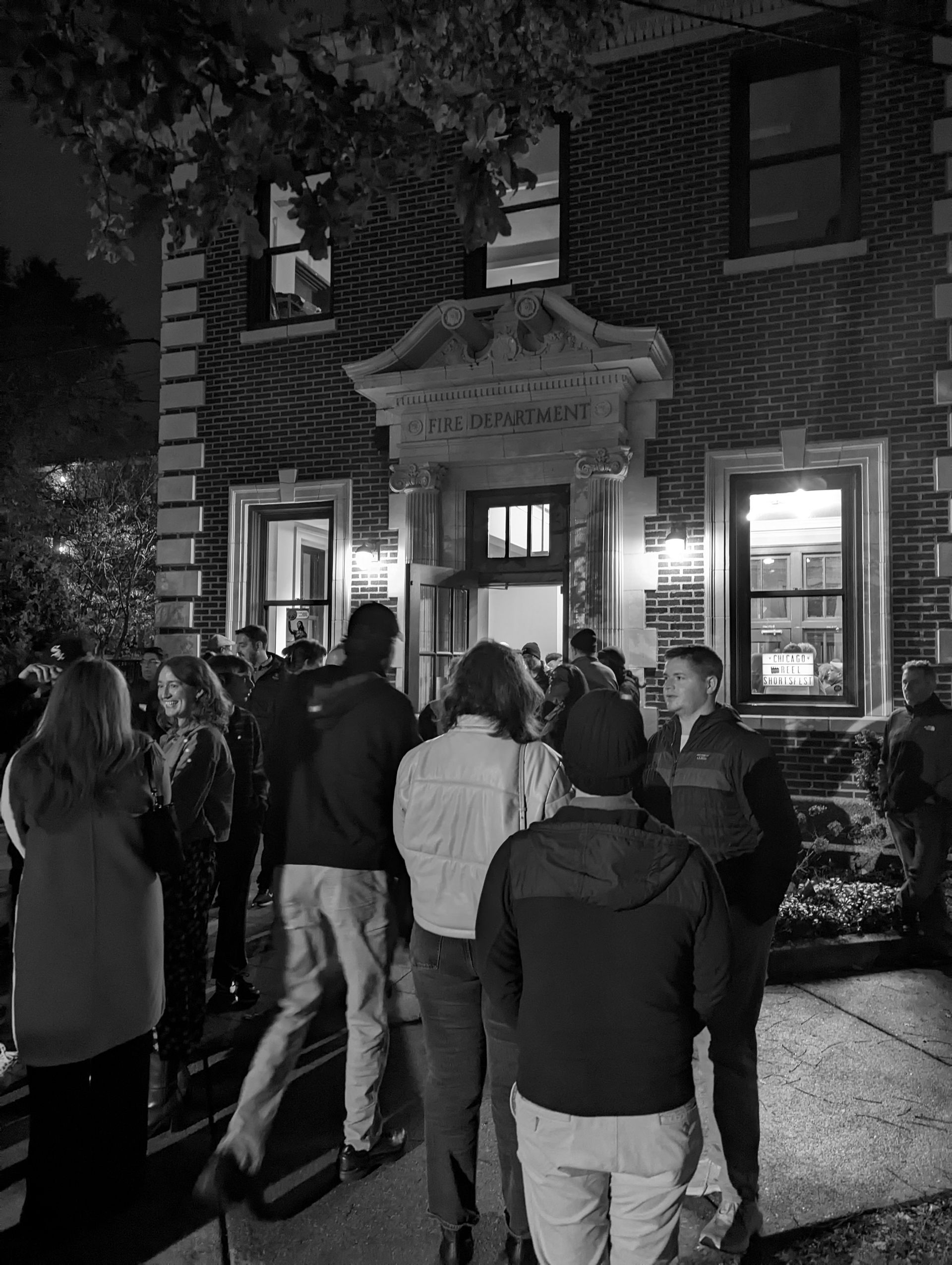 People gathered in front of a brick building at night, illuminated by outdoor lights, entering.