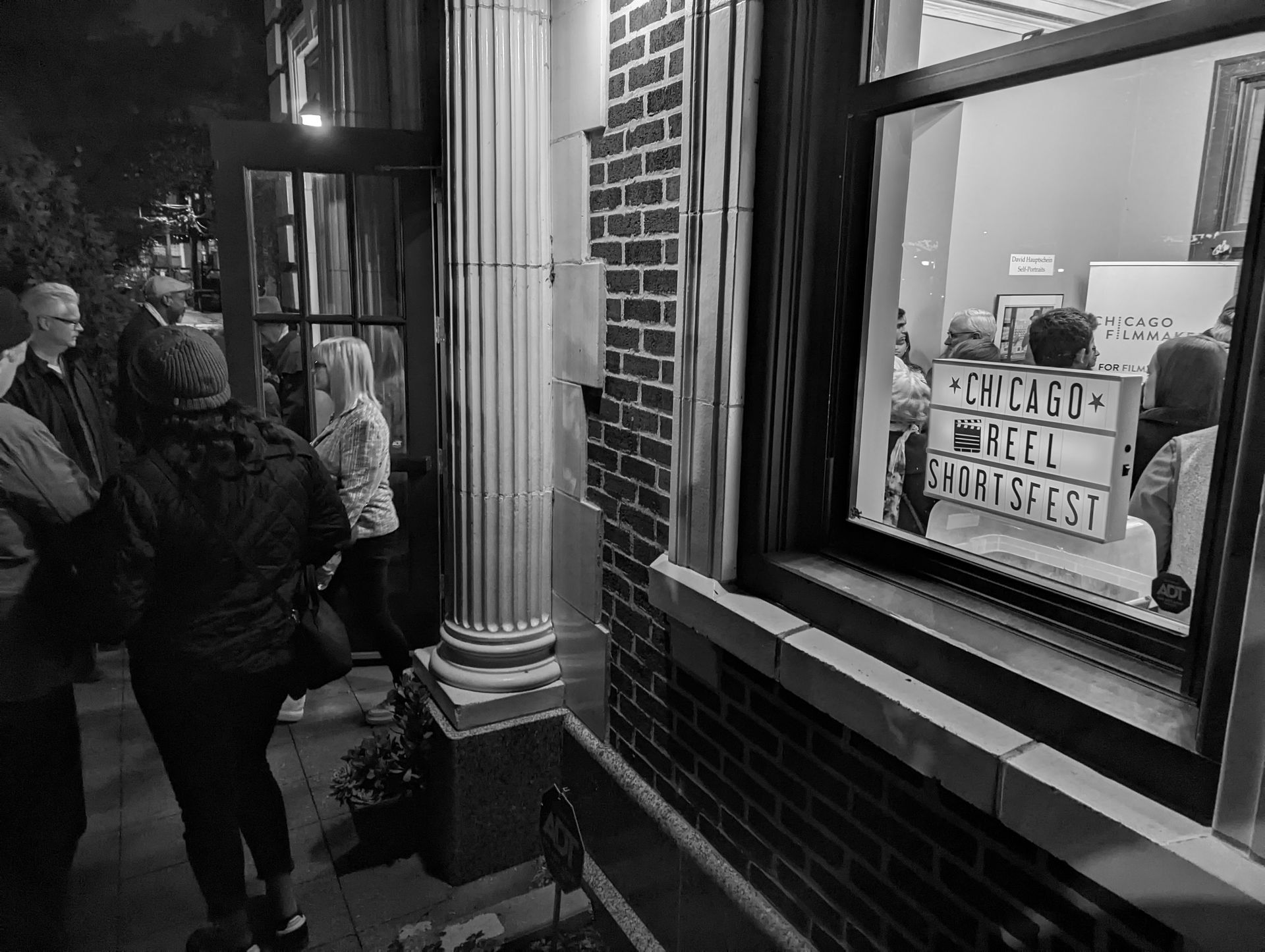 People gather outside a building. A window displays a lit sign that says 