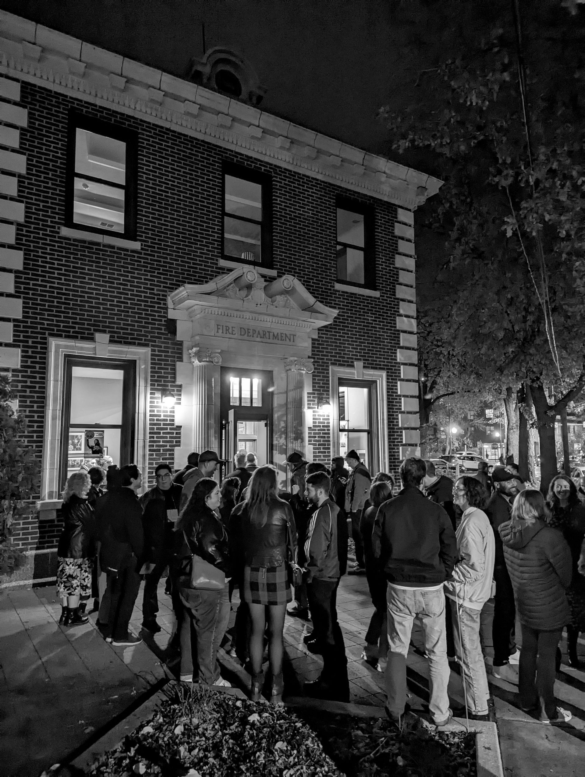 Exterior of a brick building at night with a crowd of people gathered in front of the entrance.