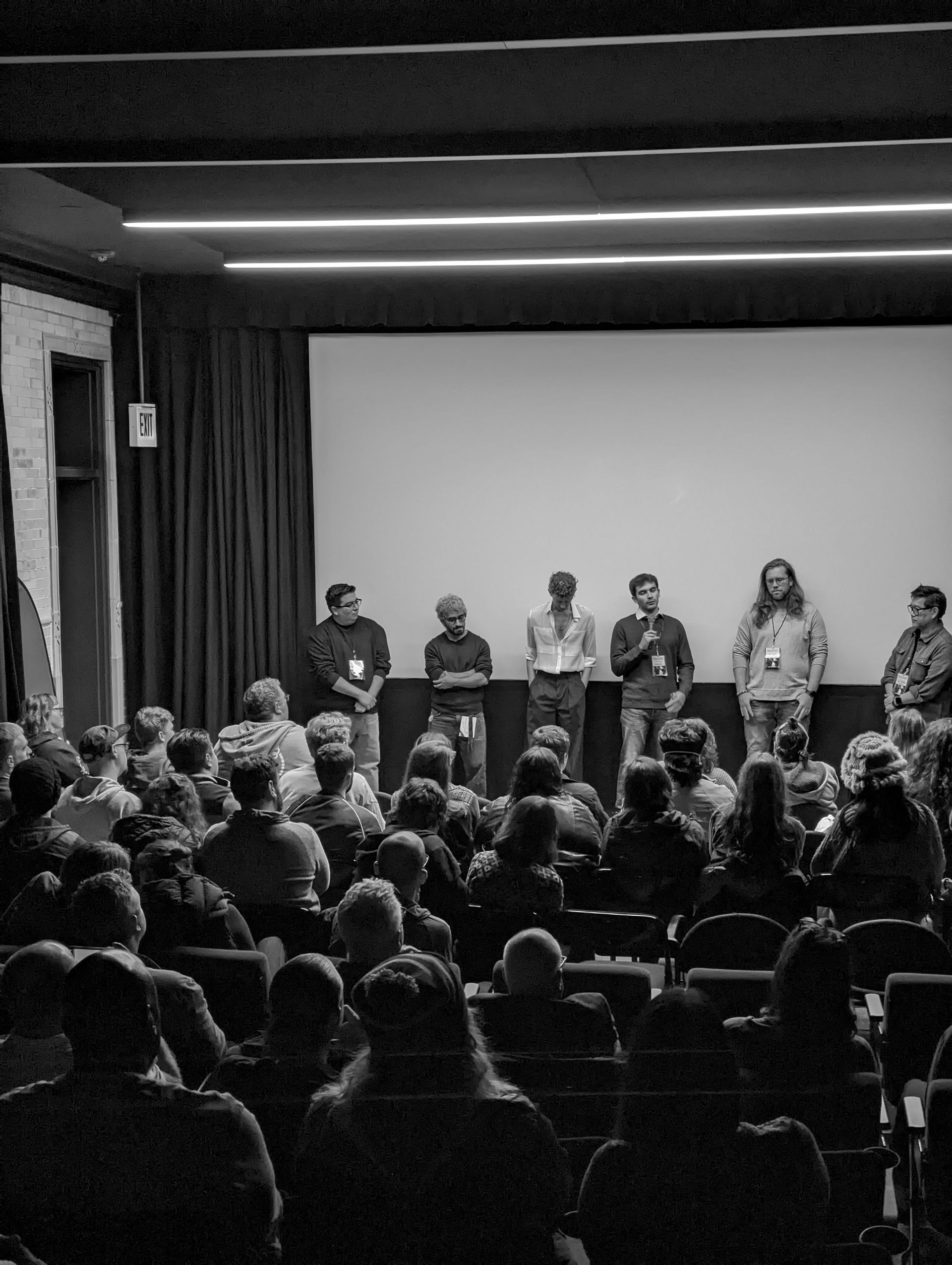 A black and white photo shows a panel of people on stage speaking to an audience in a cinema.
