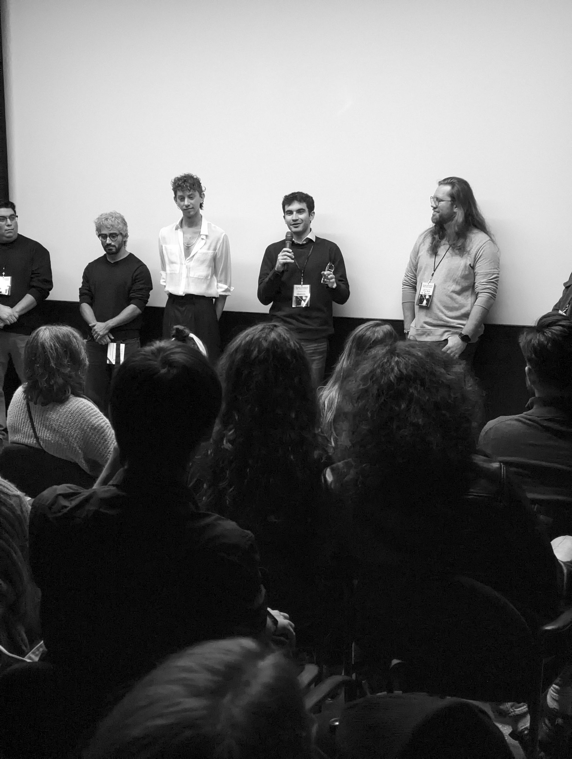 Panel of speakers in front of a screen, addressing an audience in a darkened room. Black and white.