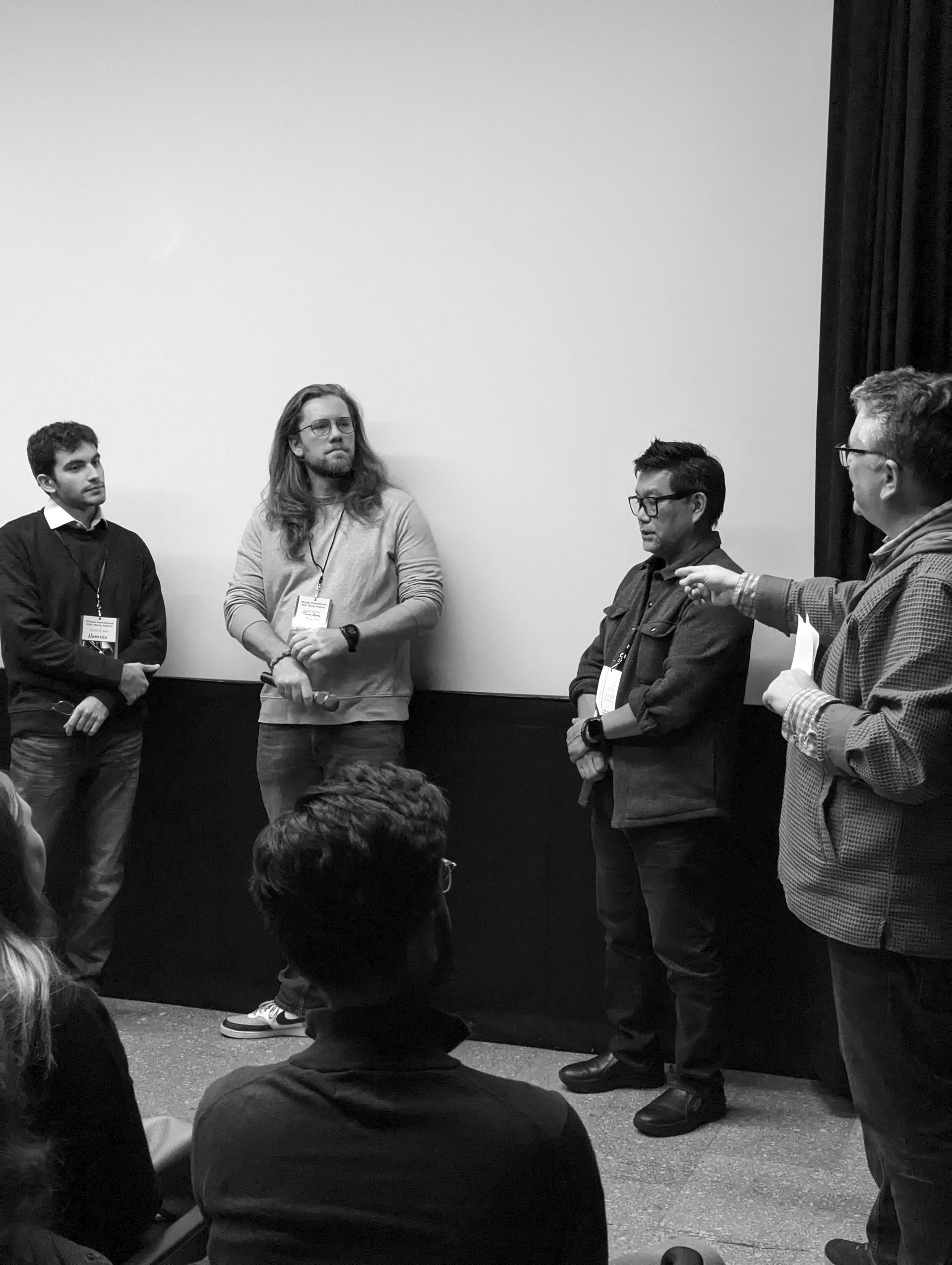 Four men standing and talking in front of a blank screen. One points while others listen.
