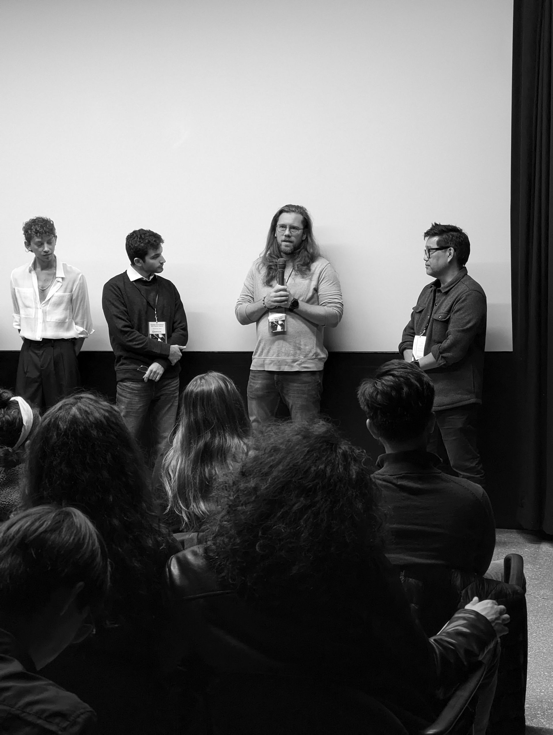 Panel of four people speaking on a stage, facing an audience in a movie theater. Black and white photo.