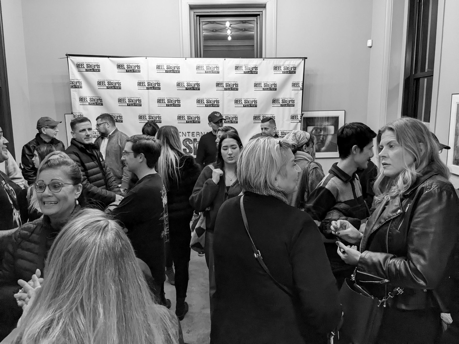Crowd at an event, socializing in a room with a banner, portraits, and neutral walls. People are engaged in conversation.