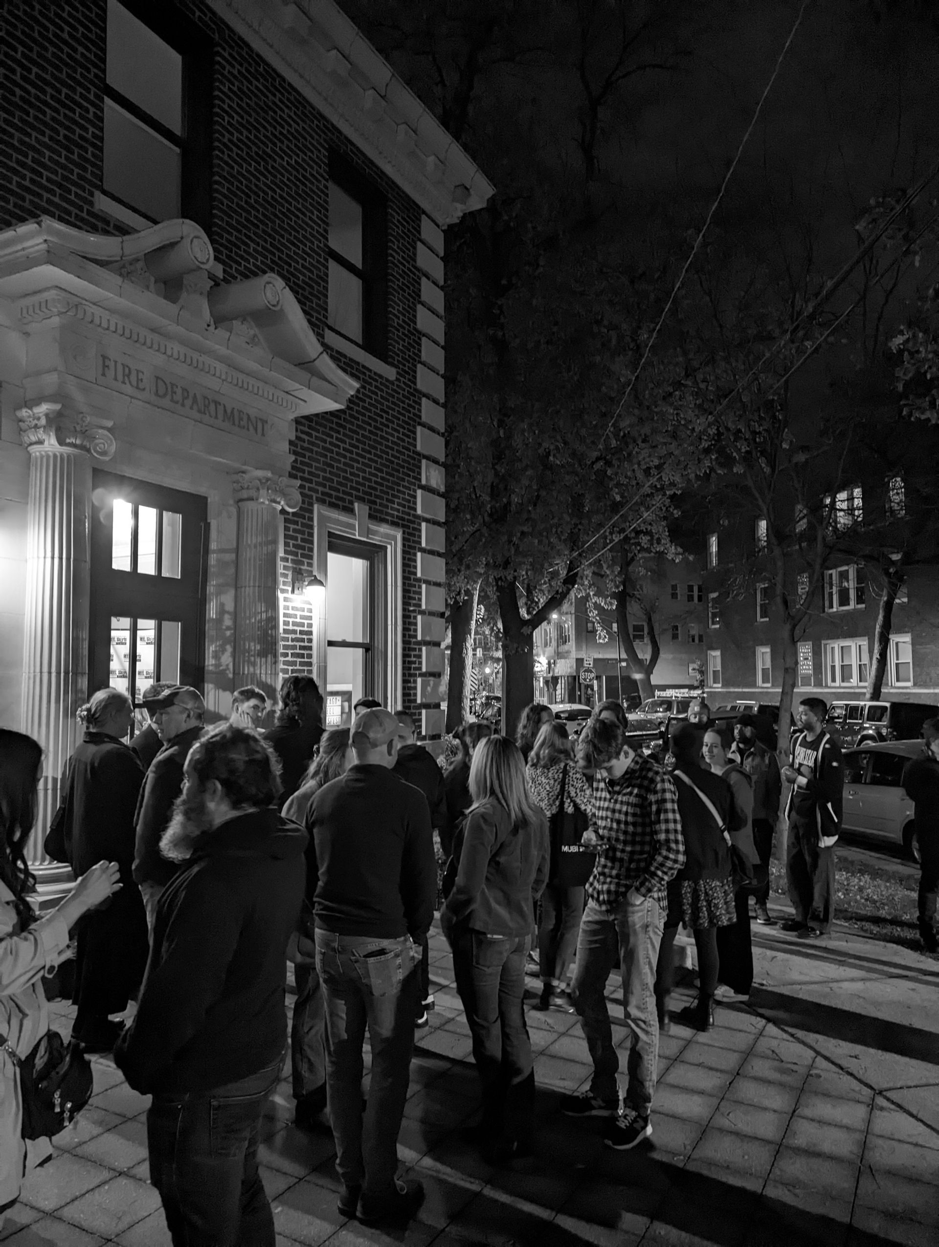 People gathered outside a brick building at night. Trees and streetlights illuminate the sidewalk.