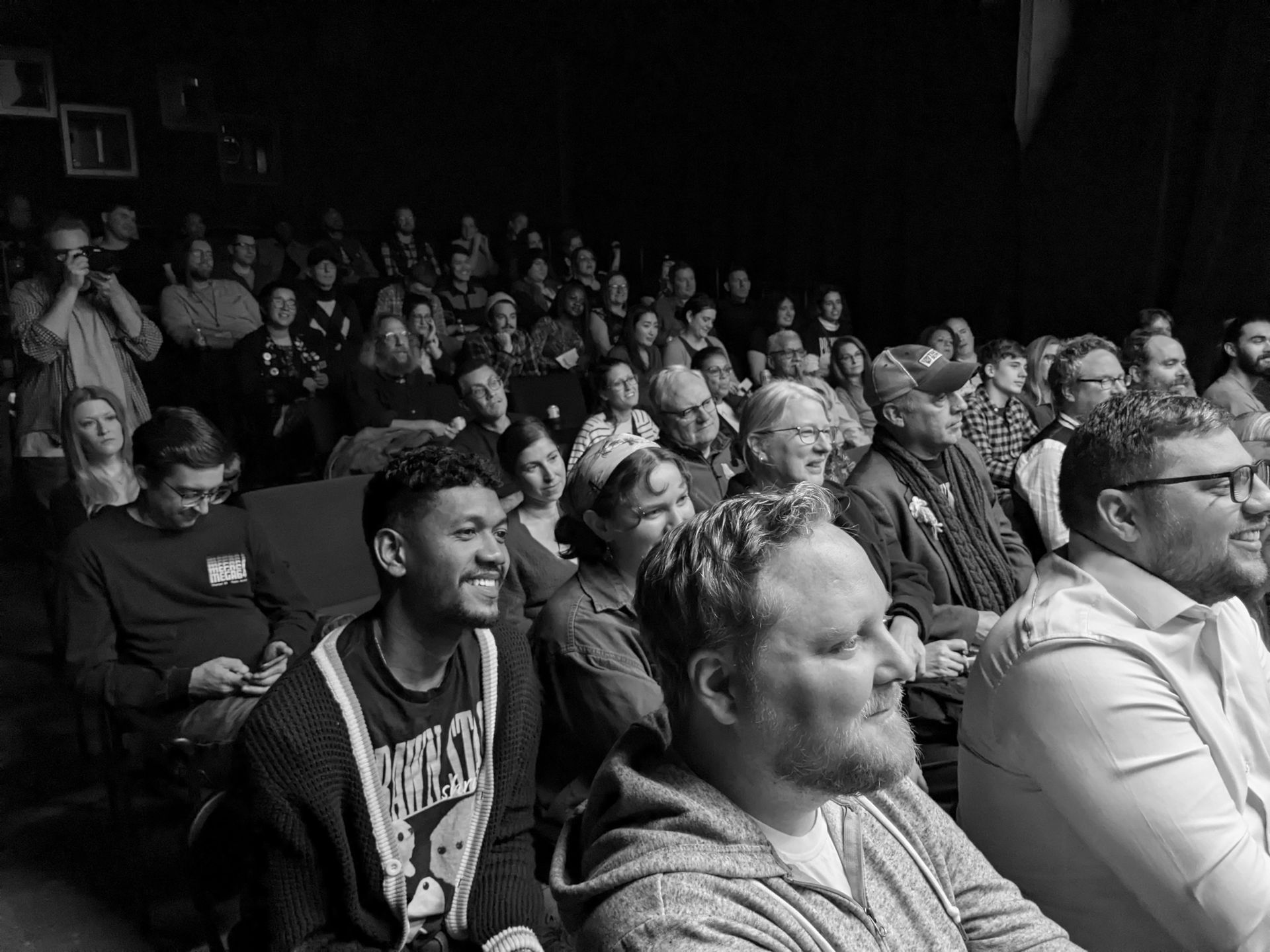 Audience in a dark theater watches a performance; many are smiling, relaxed.