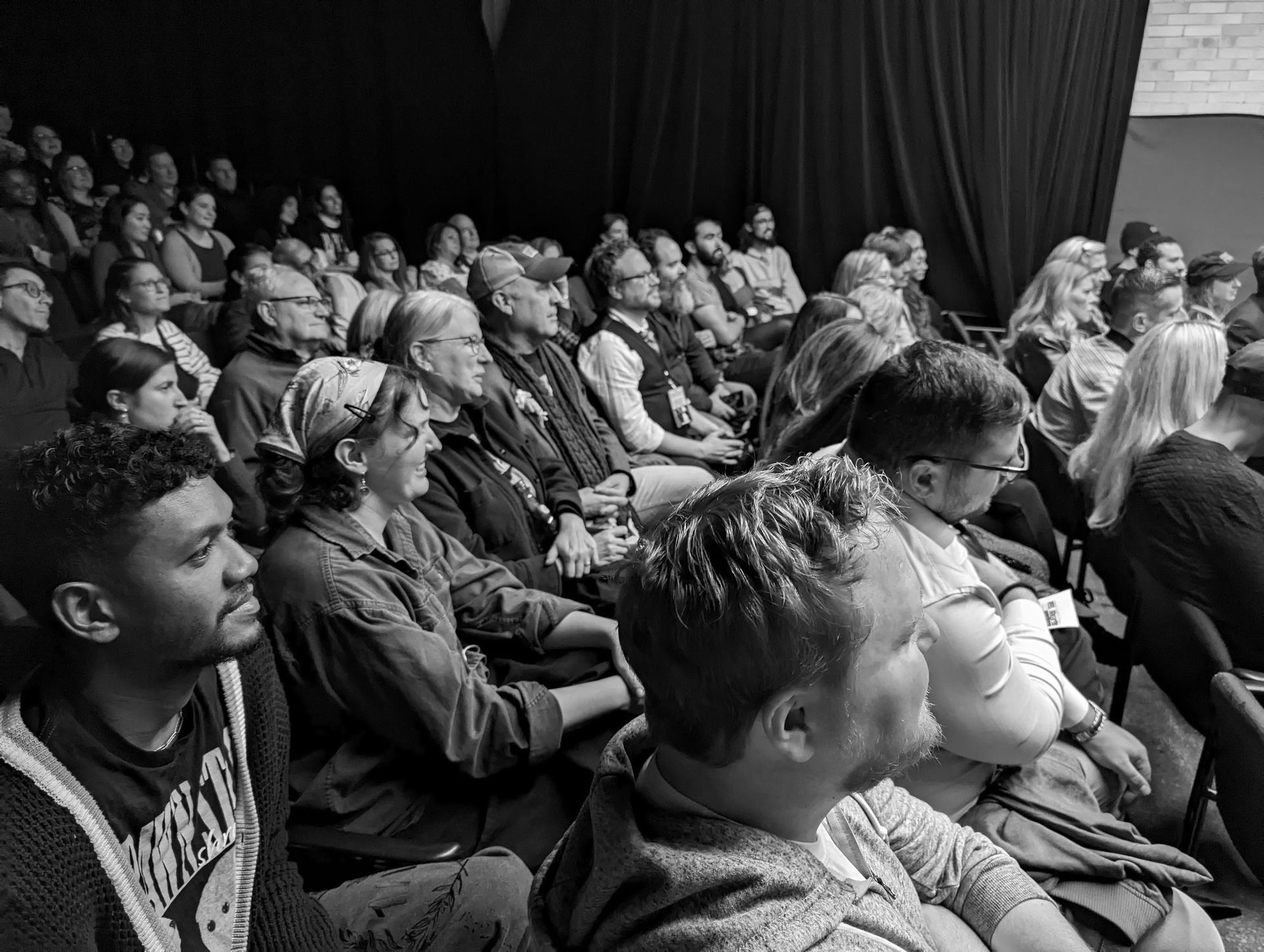 Audience in a theater, watching a performance. People of various ages and ethnicities are seated and engaged.