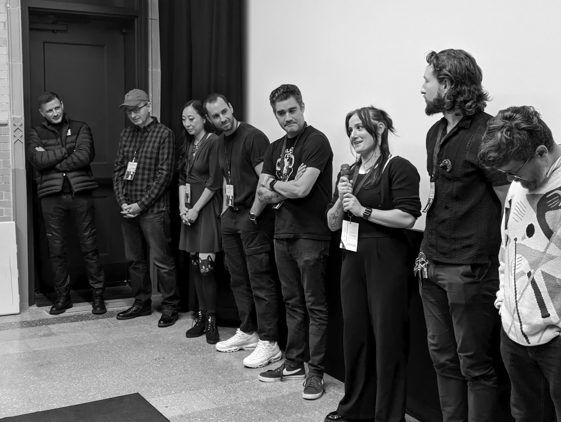 Group of people standing, some looking at the camera, one speaking into a microphone, indoors, black and white.