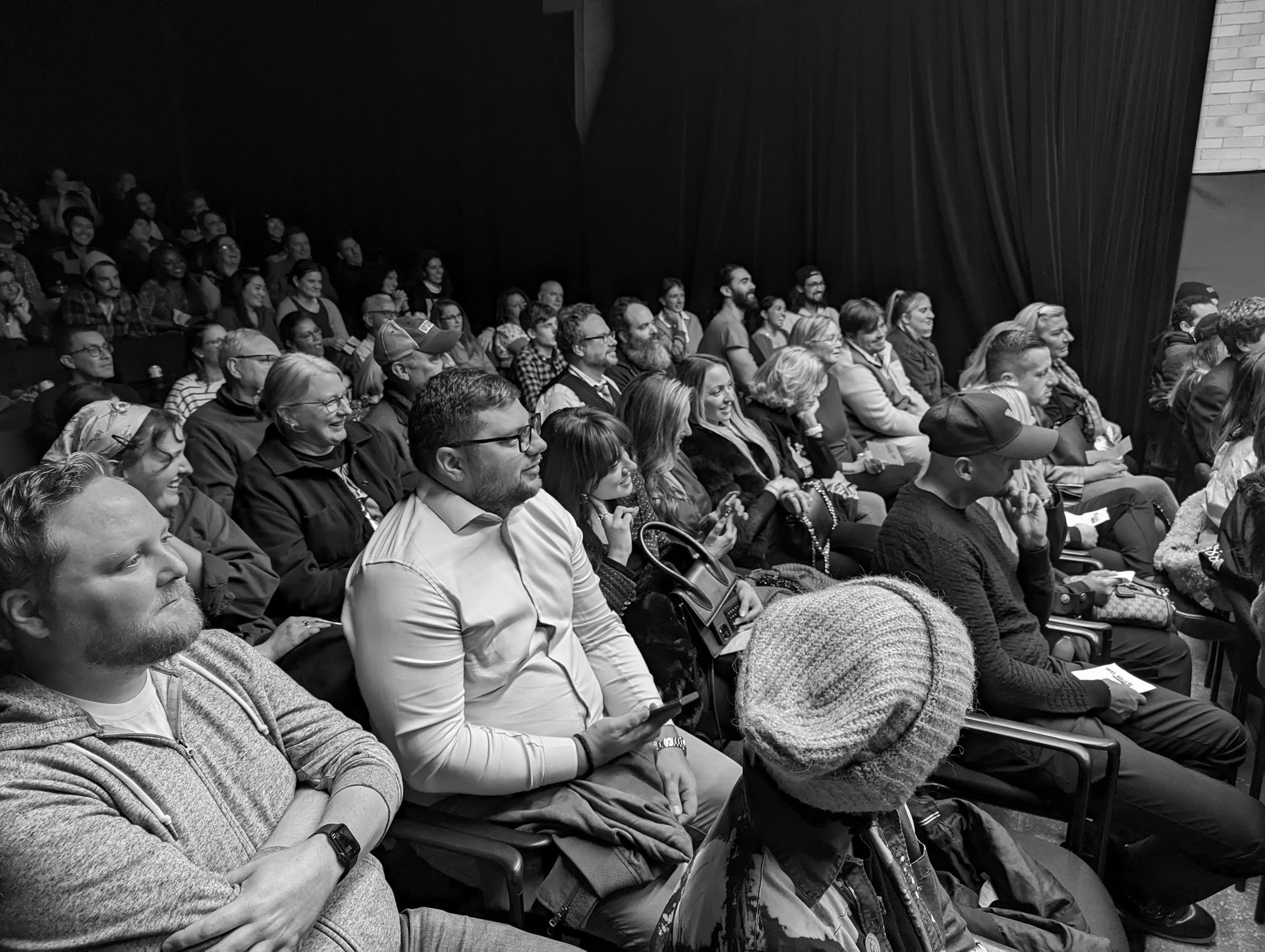 Audience in an auditorium, mostly seated, watching a performance. Men and women, mixed ages, attentive expressions.