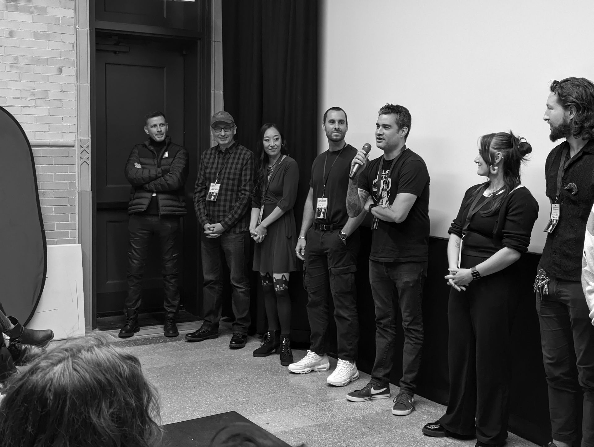 Group of people on stage, facing an audience. Black and white photo, indoor setting.