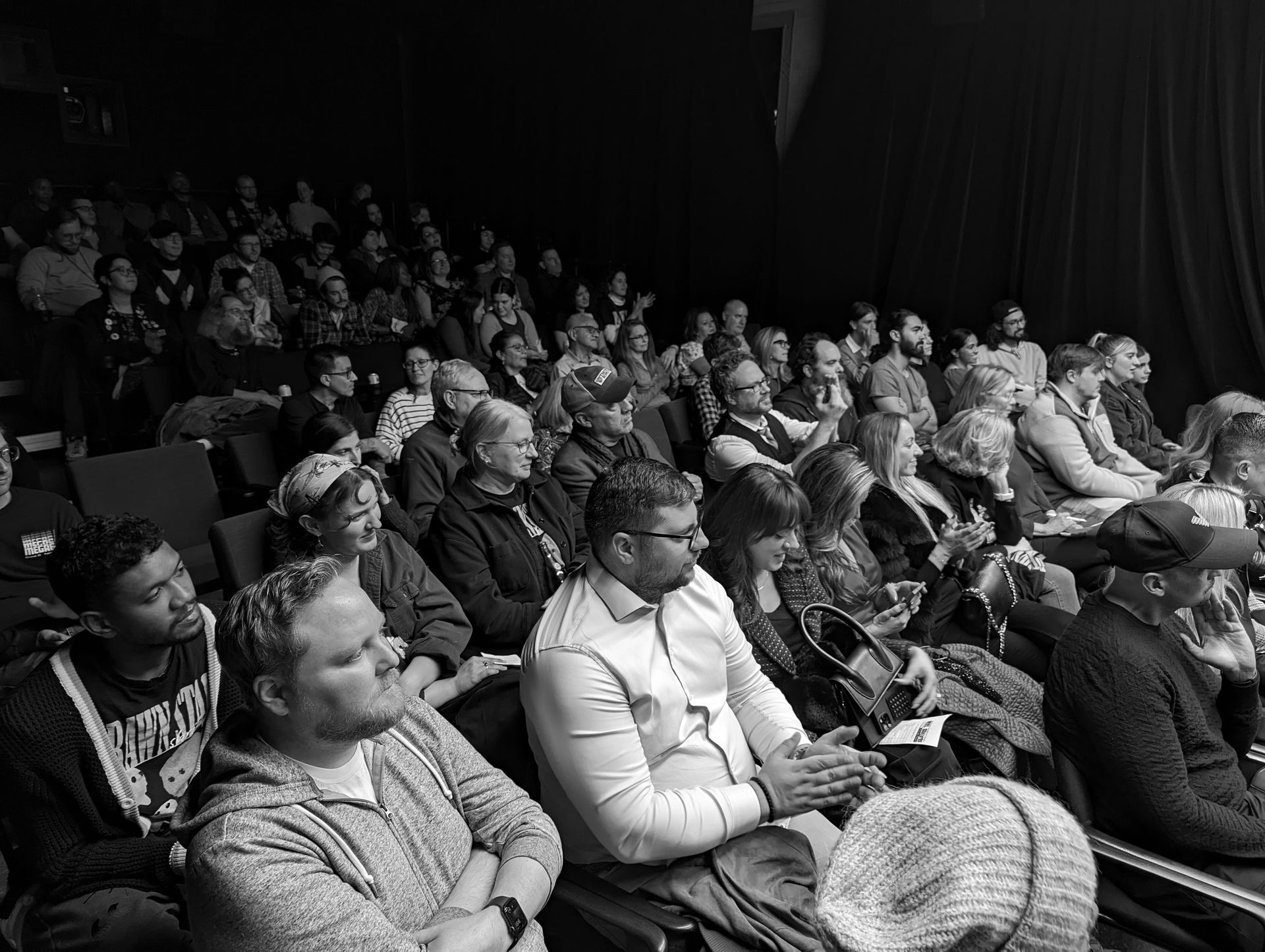Audience in theater seats; many people watching a performance. Black and white photo.